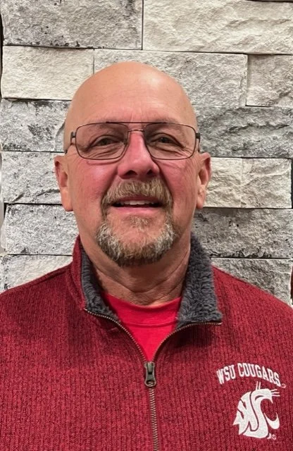 A man with glasses and a goatee wearing a red sweater with a WSU Cougars logo standing in front of a stone wall.