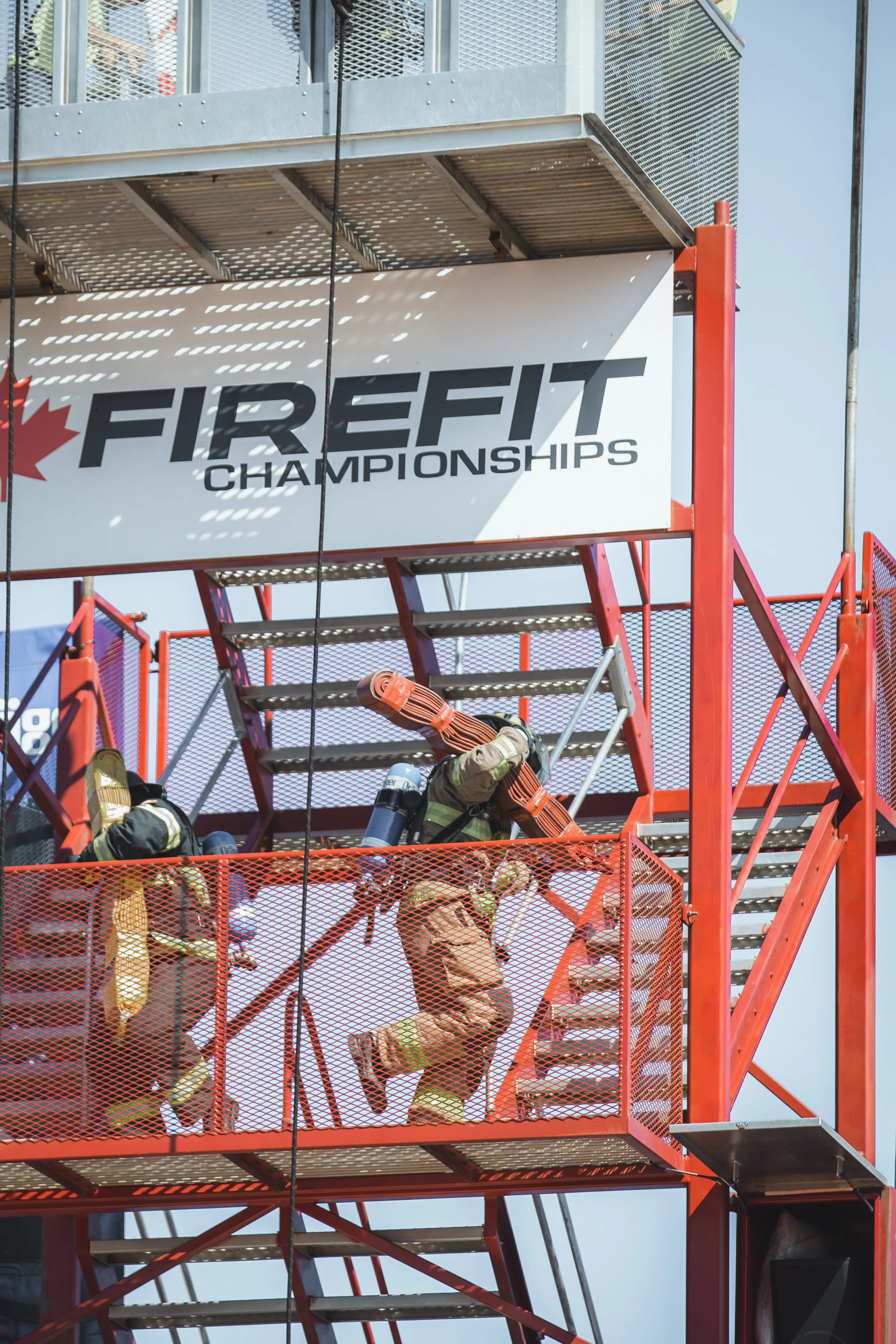 Firefighters wearing protective gear ascending a red metal staircase at a fire station during an emergency drill or competition, with a large sign reading 'FIRE FIT CHAMPIONSHIPS' and a blue sky in the background.