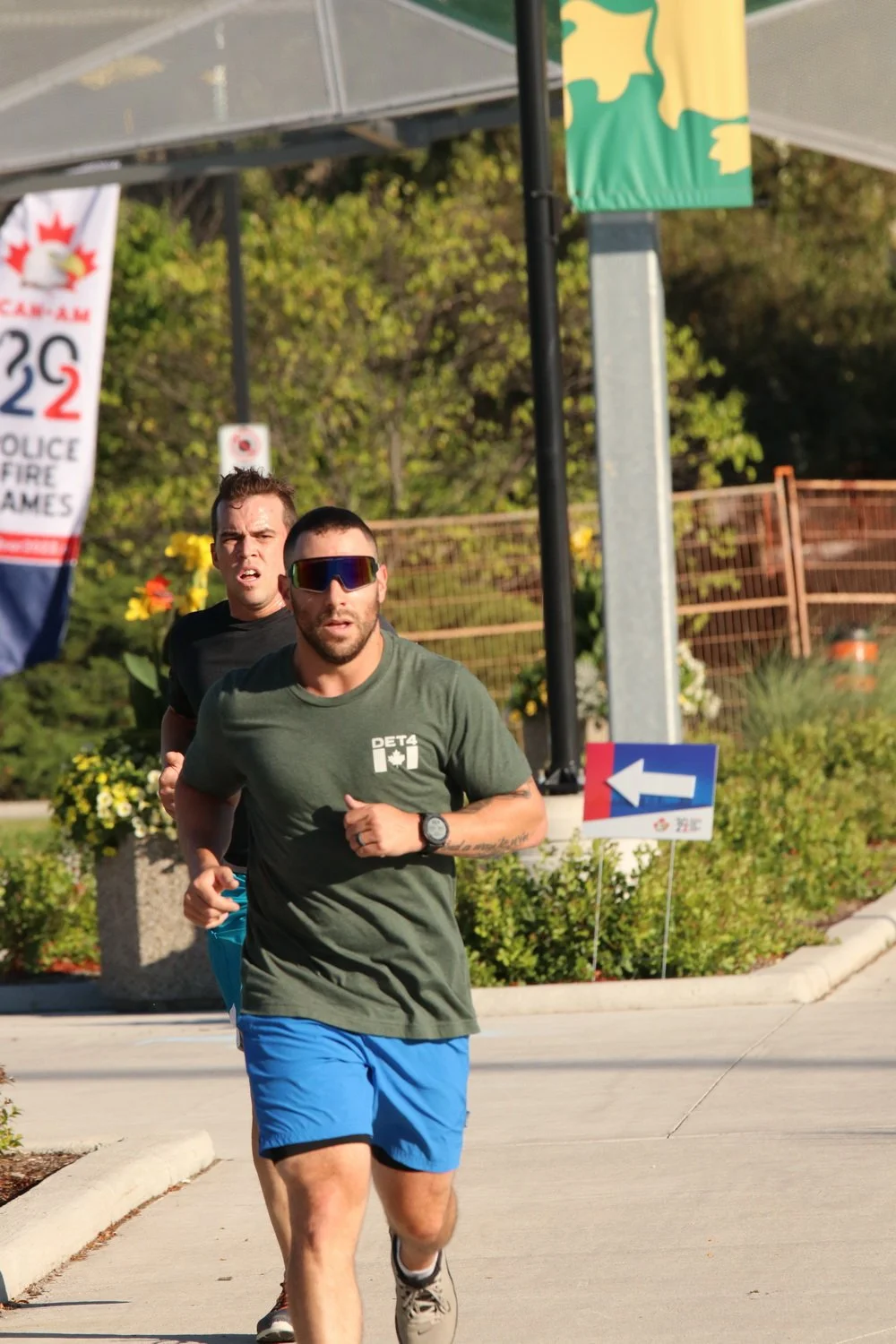 Two men running outdoors during daytime, with greenery and signage in the background.