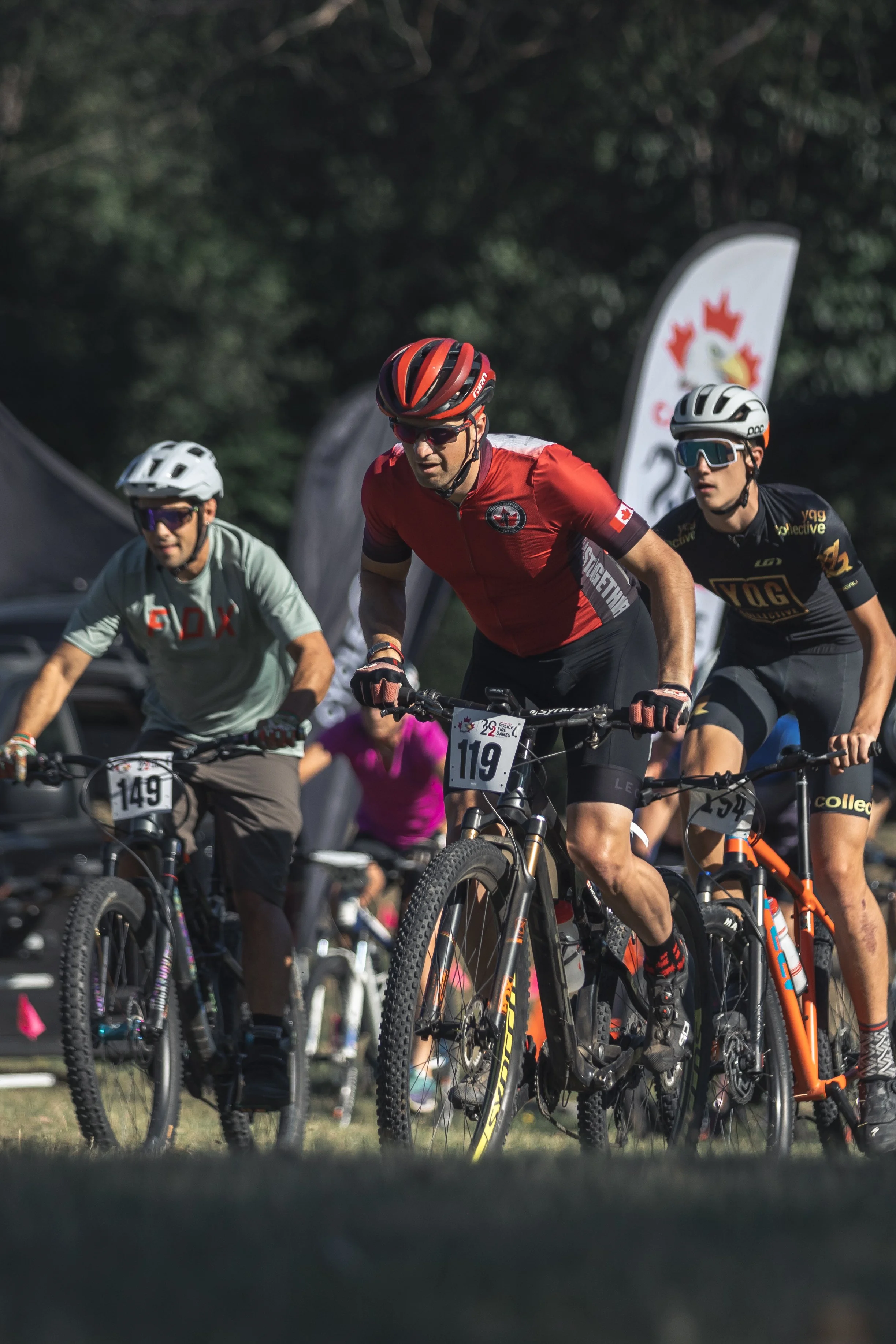 Group of cyclists participating in a race, wearing helmets and sports gear, riding mountain bikes on a trail, with a Canadian flag banner in the background.