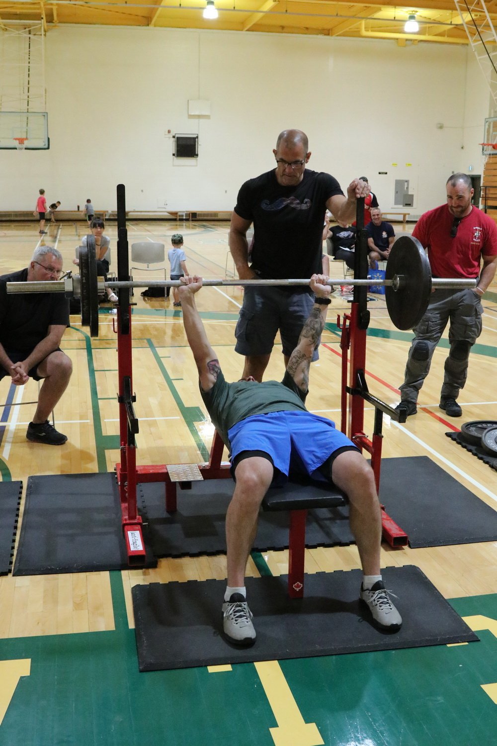 A man is bench pressing weight in a gymnasium with people around, including a spotter and several people in the background.