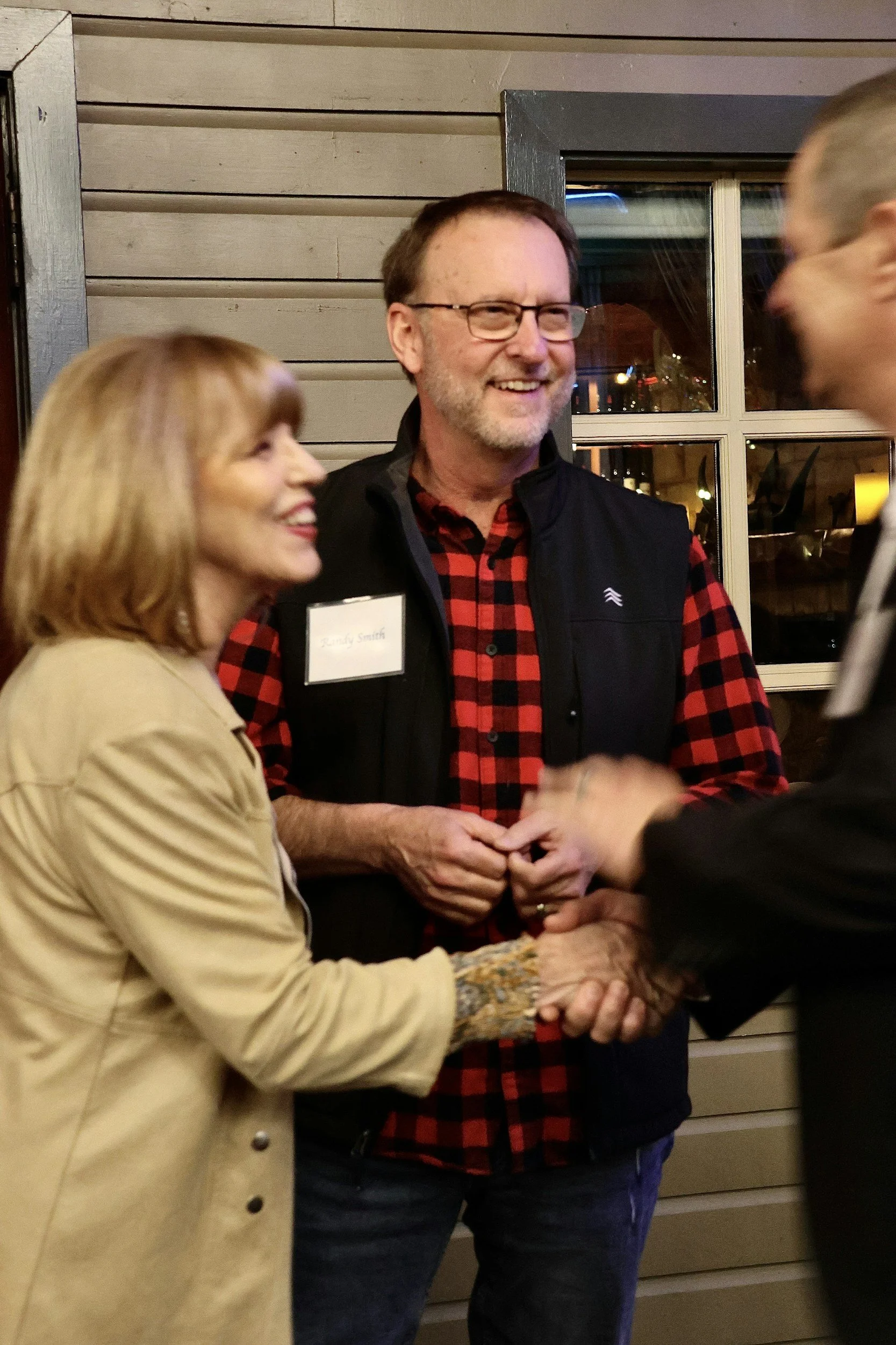 A middle-aged man with glasses and a beard wearing a red and black plaid shirt and a black vest is smiling while talking to a woman and a man at a social gathering, with a window and wooden wall in the background.