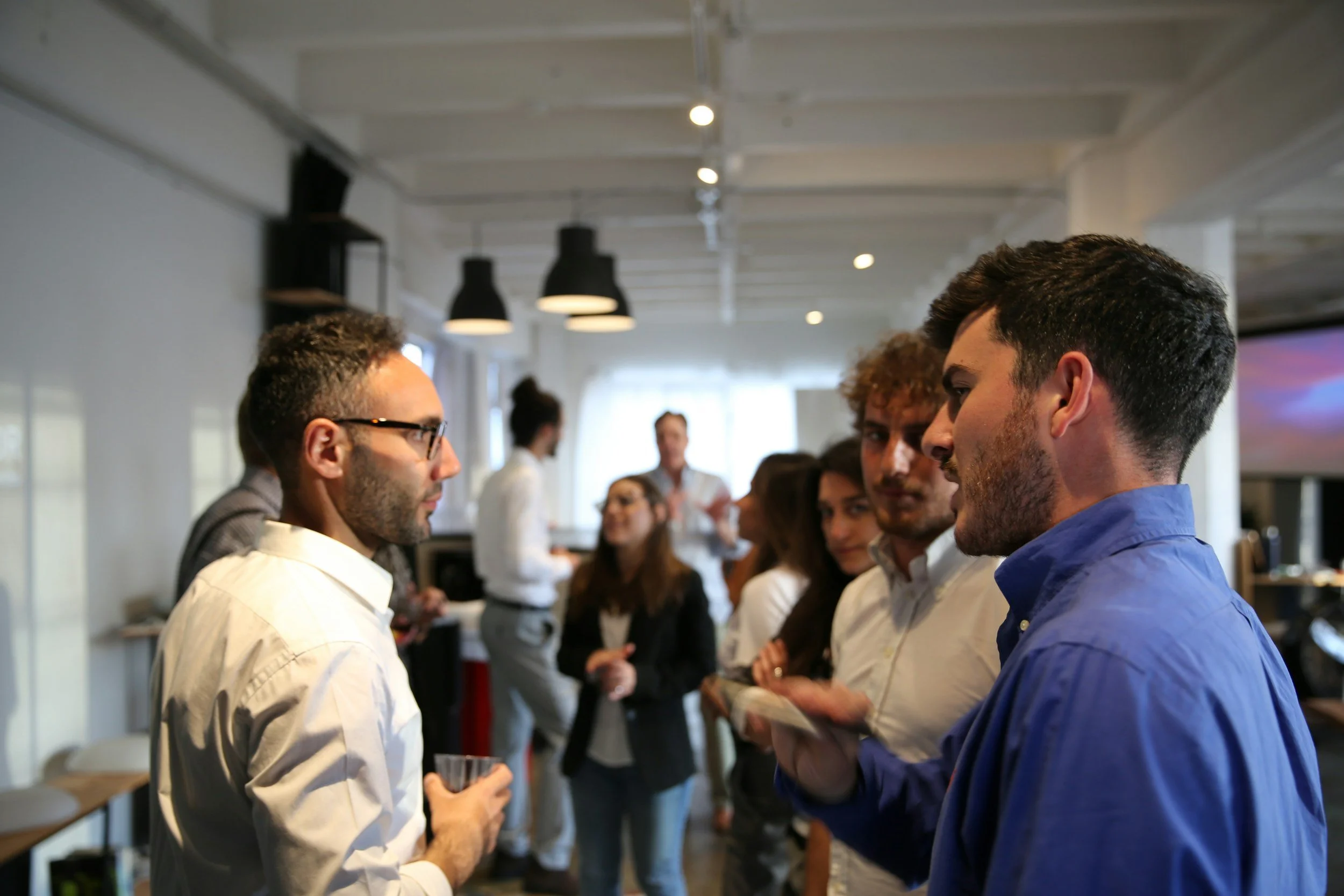 A group of people are gathered in a modern office space, engaged in conversation and networking, with some holding drinks. The environment has bright lighting and contemporary decor.
