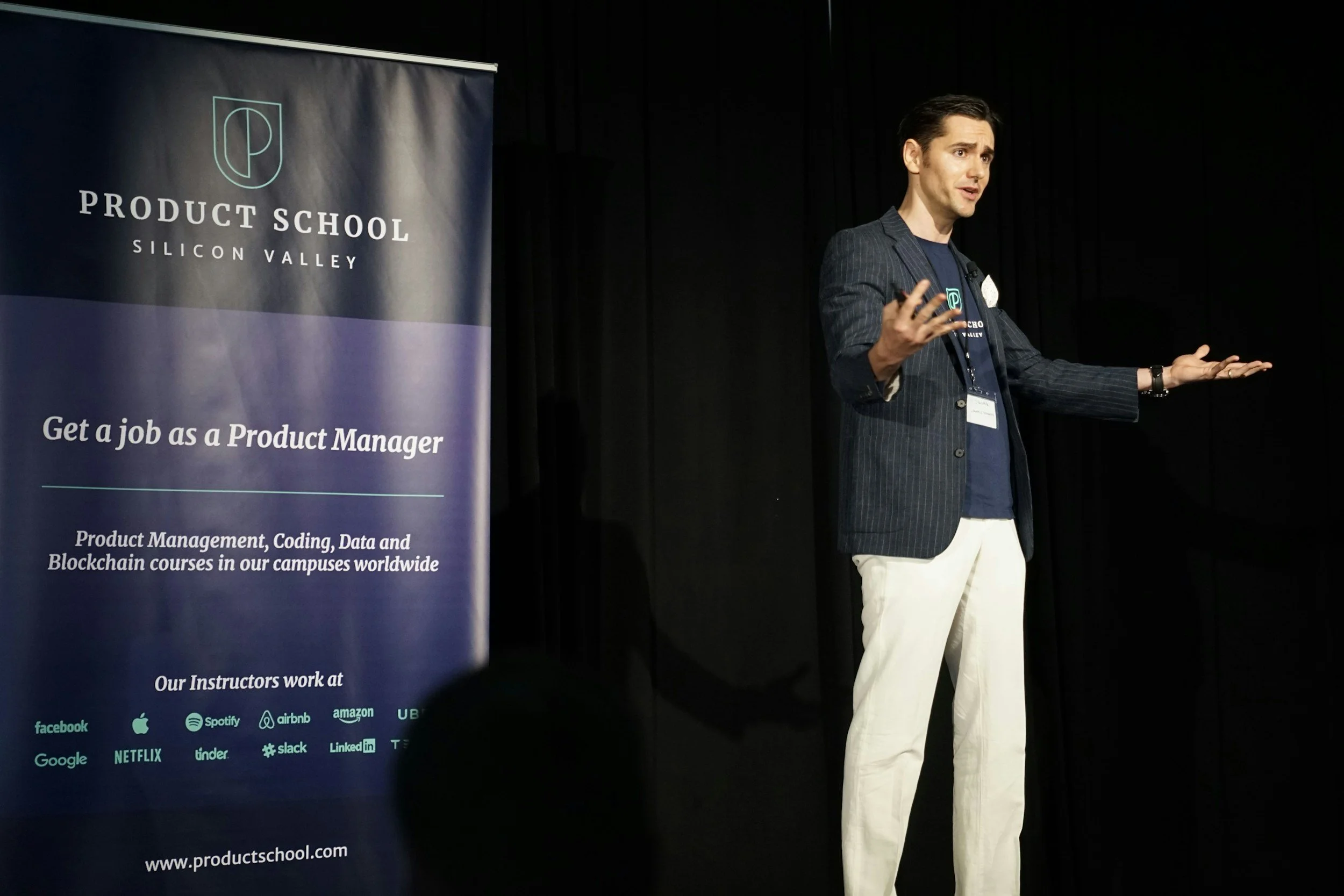 A man in a dark pinstripe blazer and white pants is speaking on stage at a Product School event in Silicon Valley. A large banner beside him promotes the Product School courses and lists companies like Facebook, Apple, Spotify, Airbnb, and others as their partners.