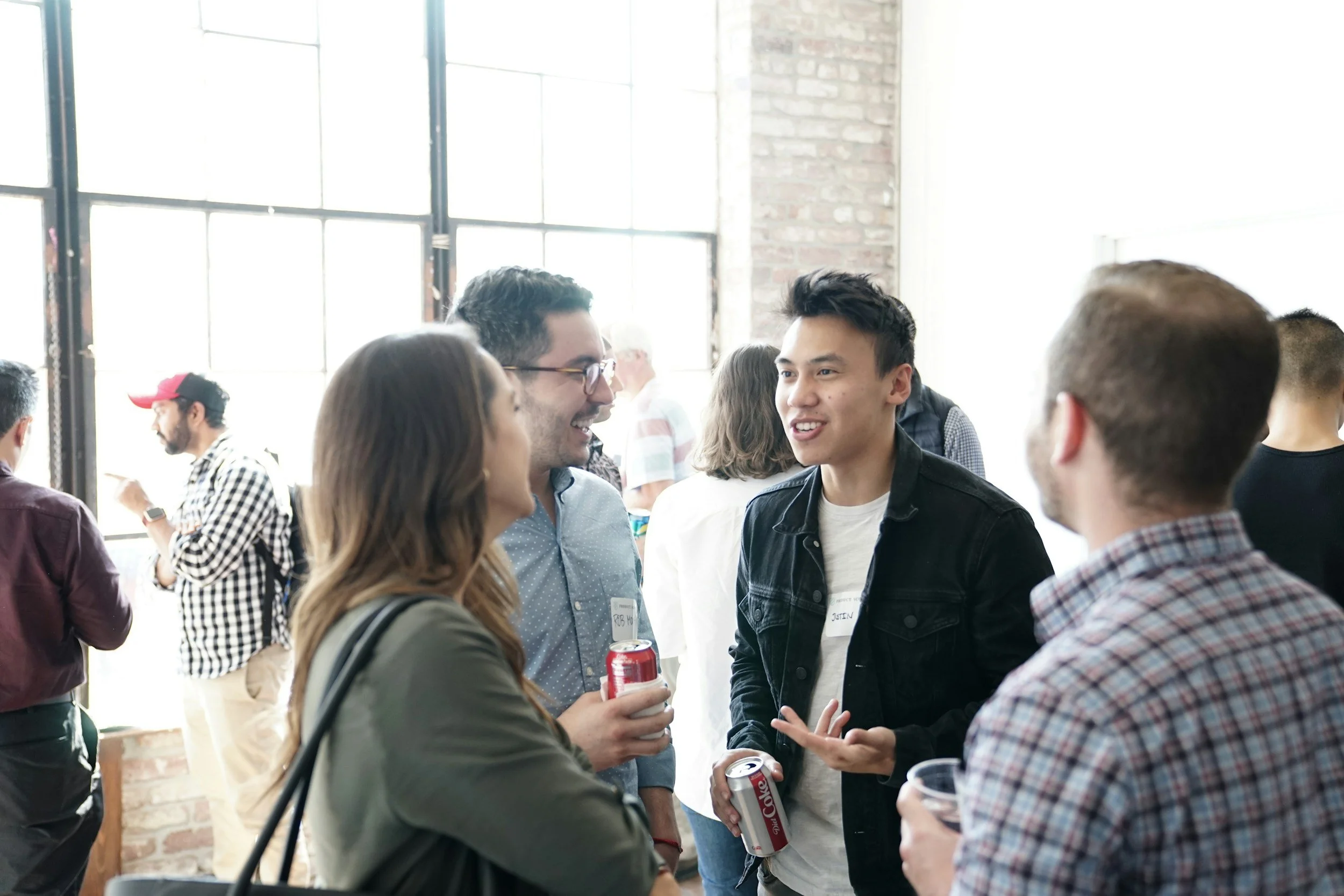 Group of people socializing at an indoor event, some holding cans of soda, in a room with large windows and brick walls.