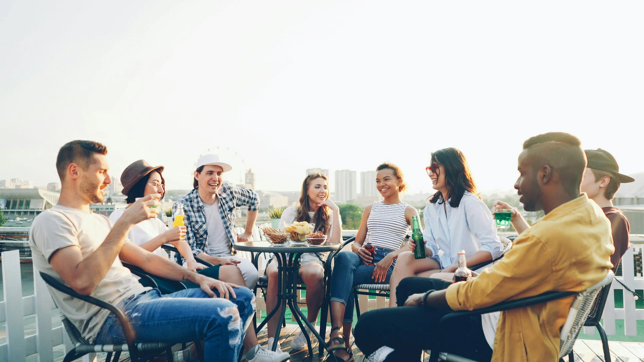 A group of friends smiling and enjoying drinks on a rooftop patio with city buildings in the background.