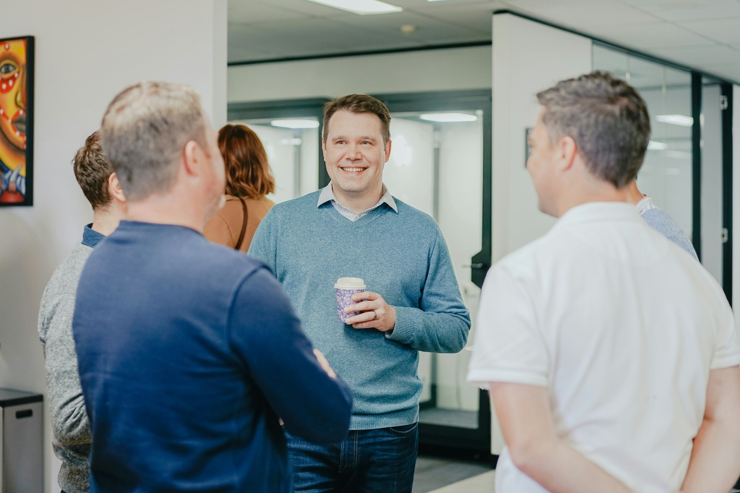 A man in a blue sweater holding a coffee cup, smiling, and talking to three other people in an office hallway.