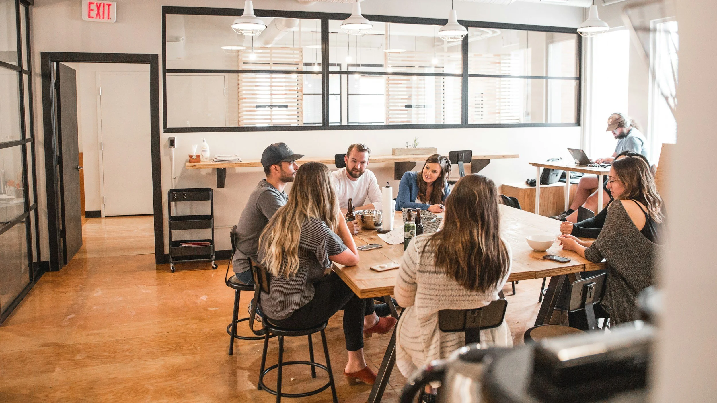 A group of eight people sitting around a large wooden table in a modern, well-lit office space, engaging in conversation.