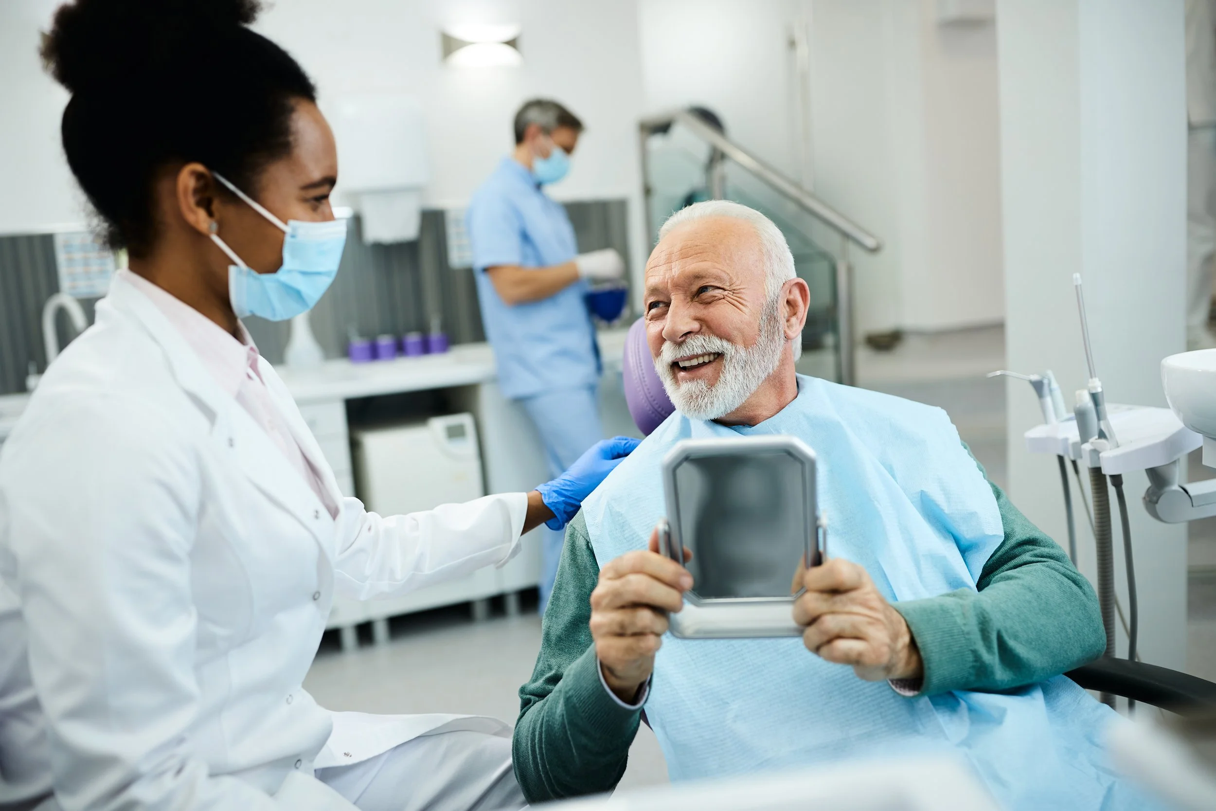 An elderly man with a white beard smiling and looking at his reflection in a mirror while sitting in a dental chair, being attended to by a female dental assistant wearing a surgical mask. Another healthcare professional in the background is working.