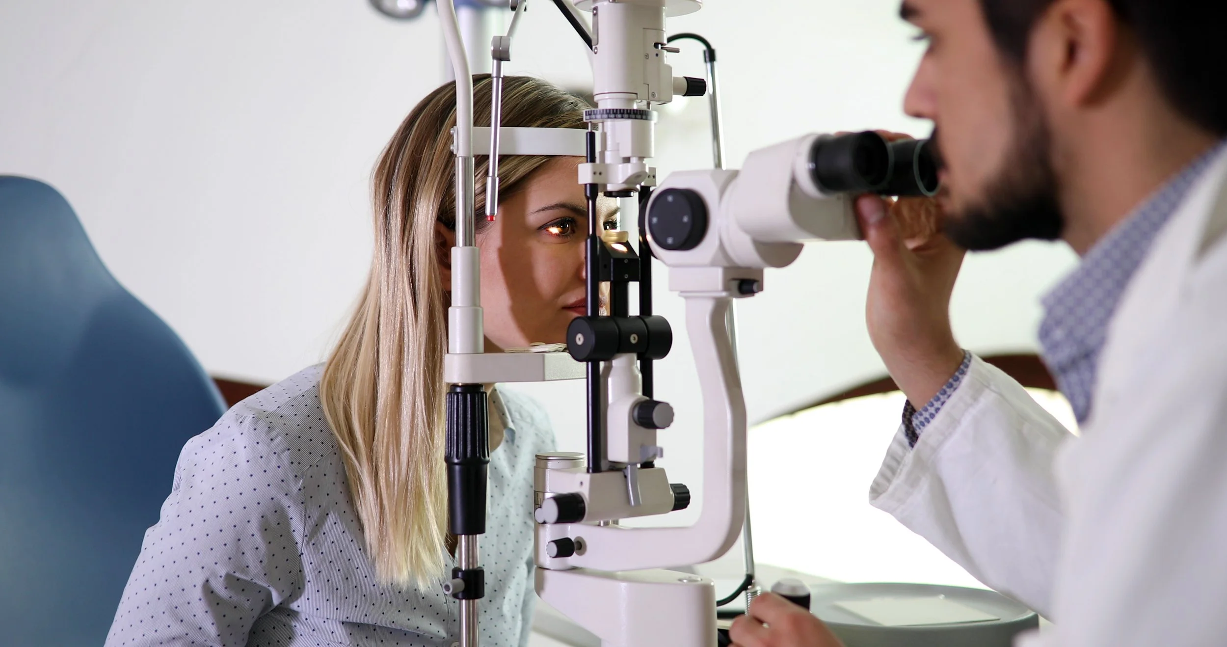 An eye examination at an optometrist's office, with a woman looking into a slit lamp and a male optometrist using a microscope.