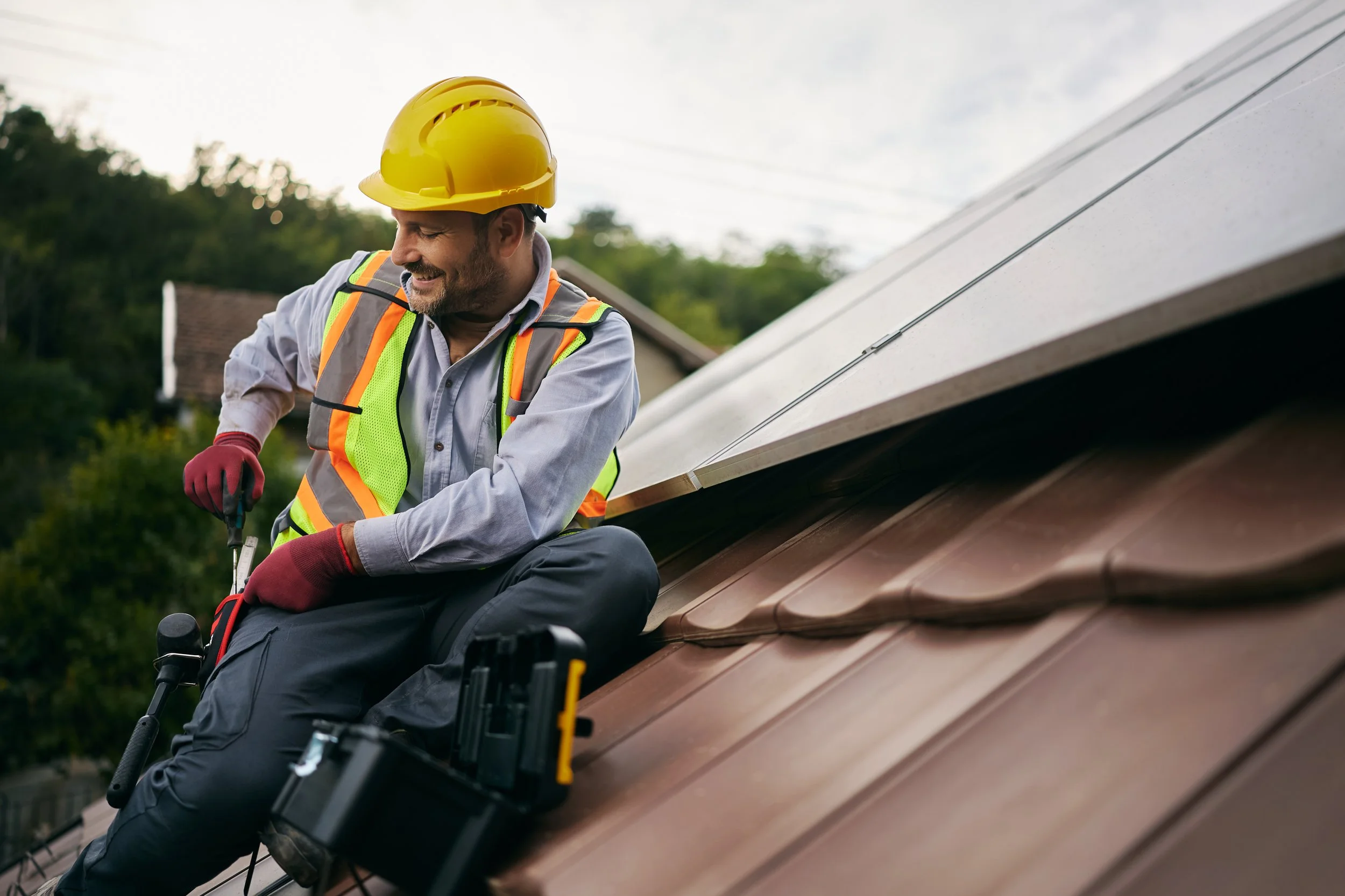 A man wearing a yellow safety helmet and a reflective vest is sitting on the edge of a roof, working with roofing tools during daytime.