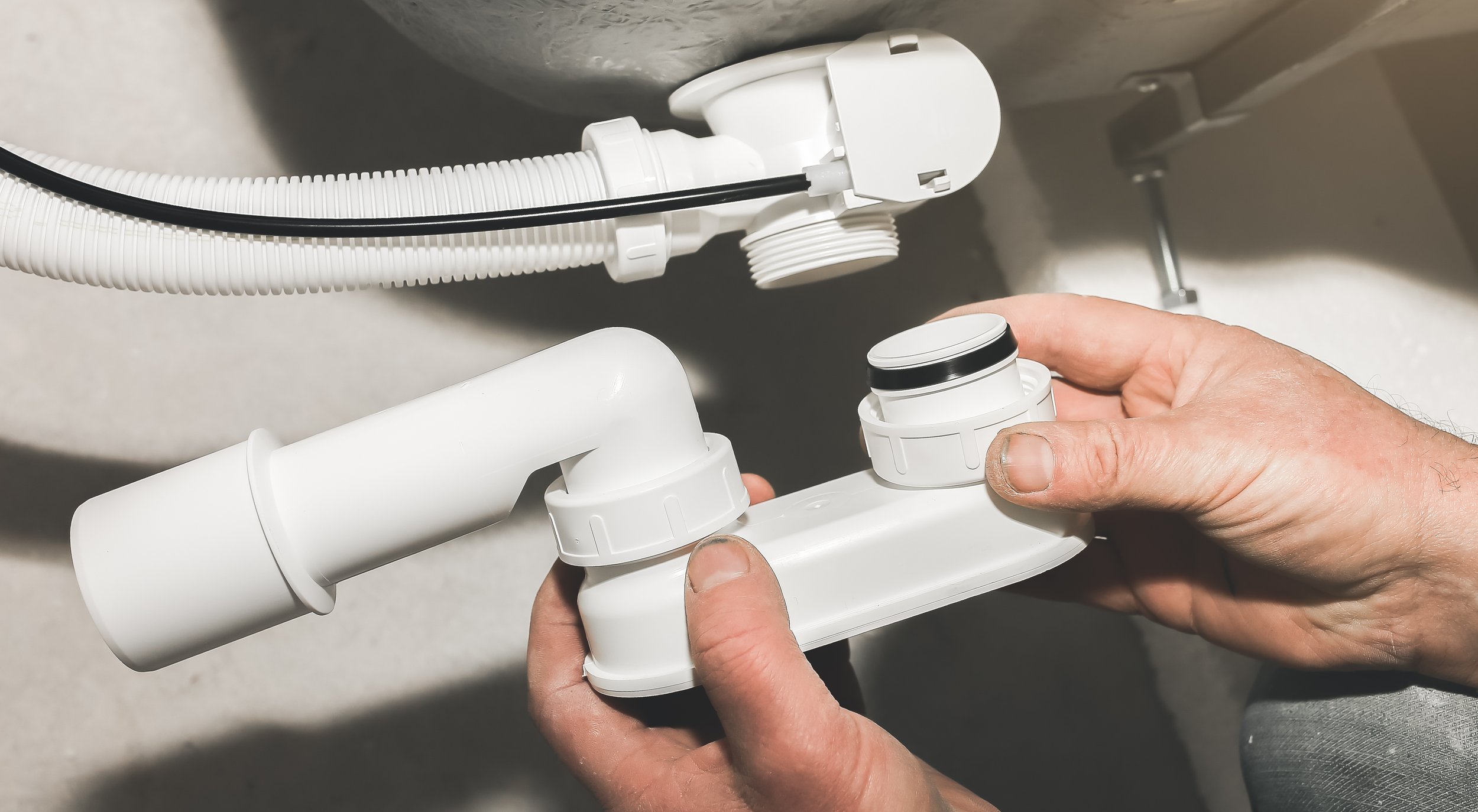 Close-up of a person adjusting plumbing connections under a sink, focusing on white PVC pipes and fittings.