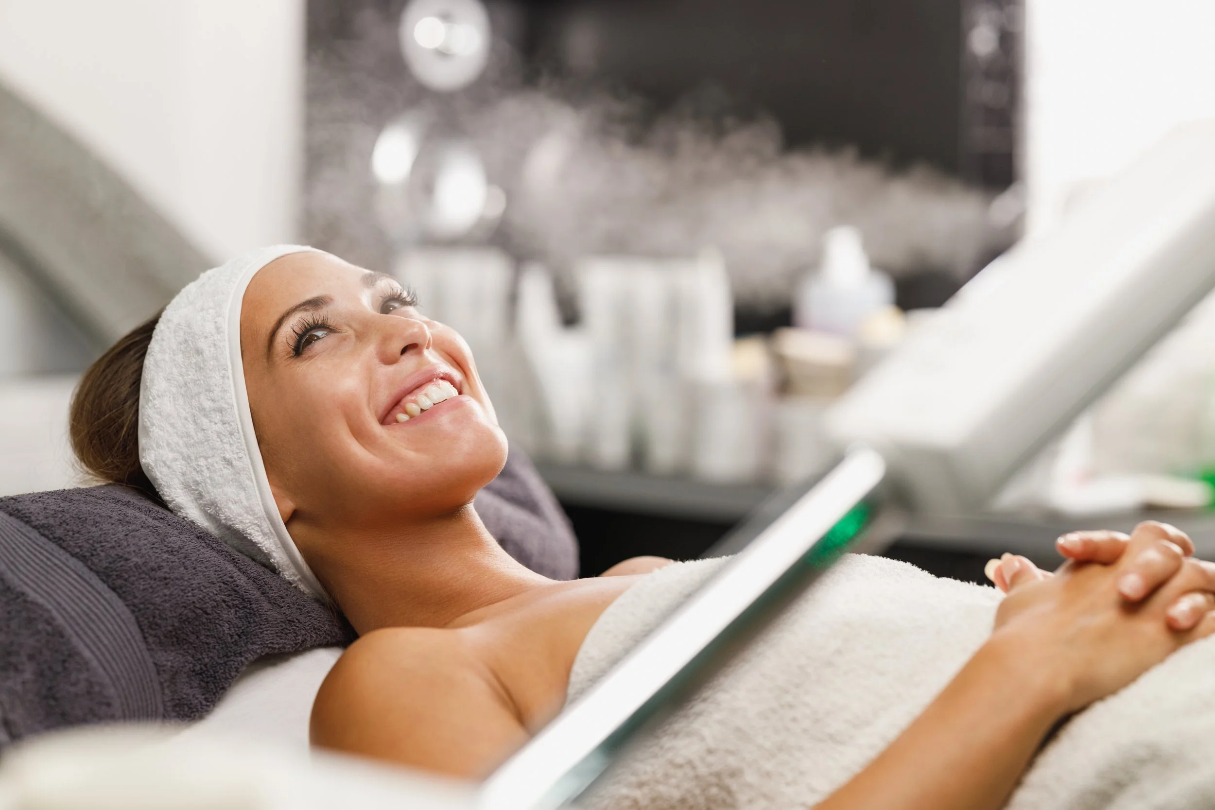 Smiling woman with towel on her head lying in spa or salon with steam in the background.