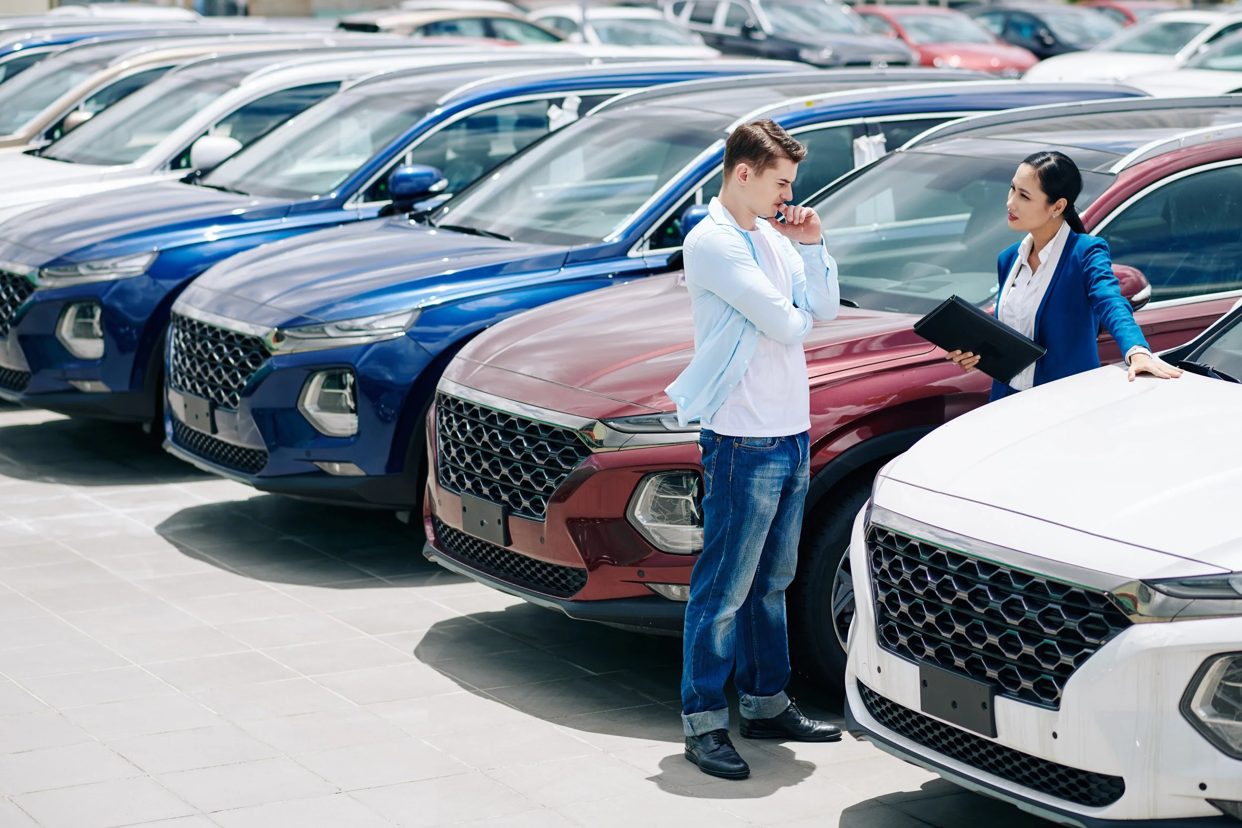 A man and woman, possibly a car salesperson and customer, standing among parked cars at a dealership, engaged in conversation.