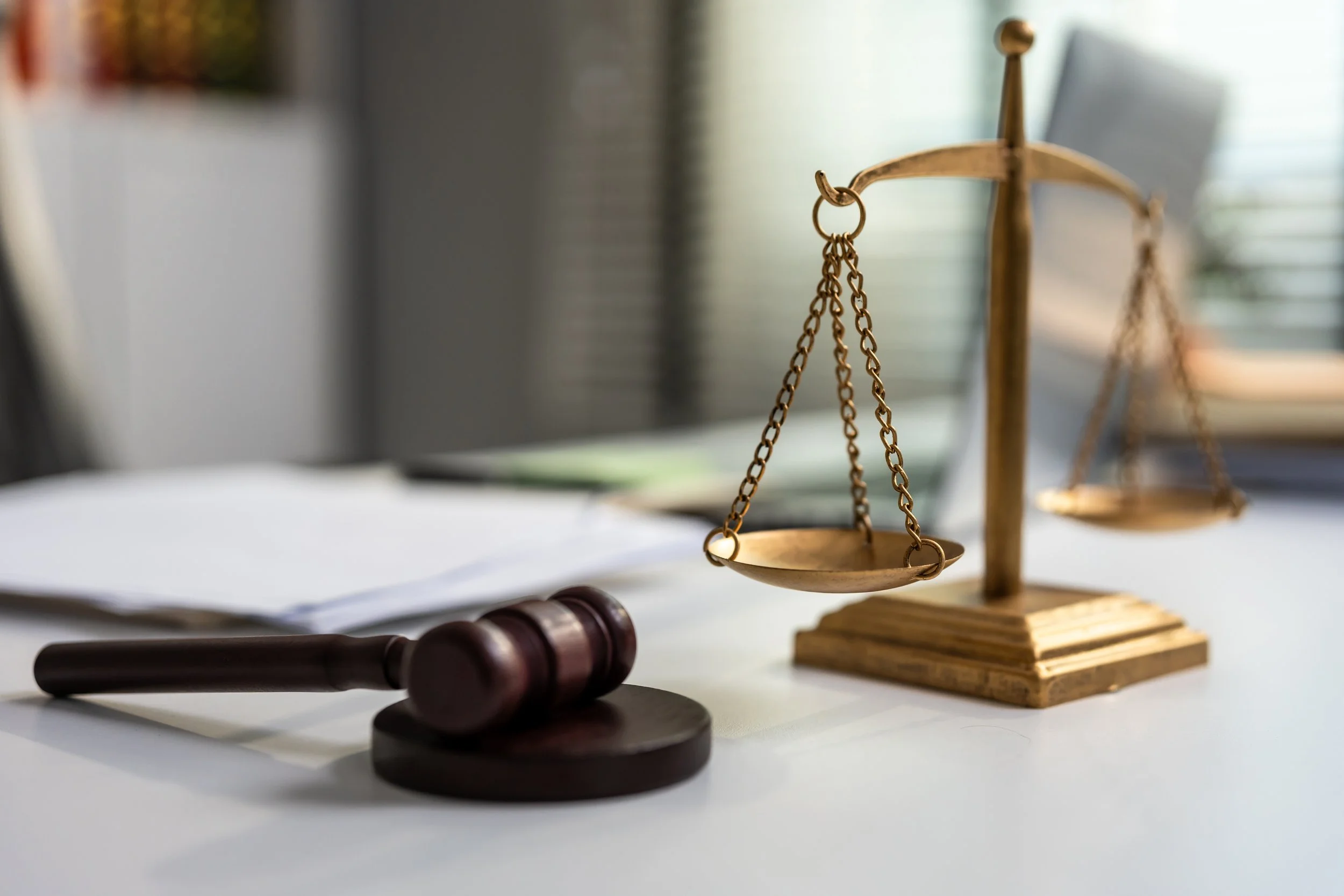 A small brass balance scale on a wooden base, placed on a white surface with papers and a gavel nearby, in a setting resembling a law office or courtroom.