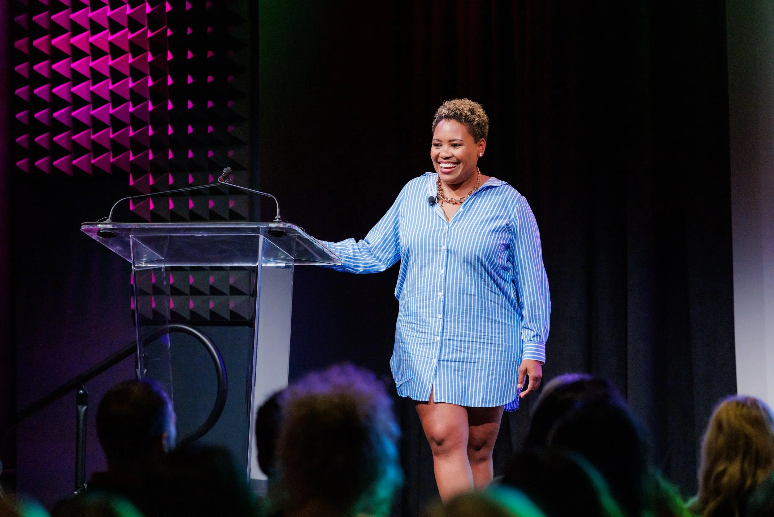 A woman standing at a clear podium on a stage, smiling, wearing a blue and white striped button-up shirt, and surrounded by an audience. There are purple and black acoustic panels behind her.
