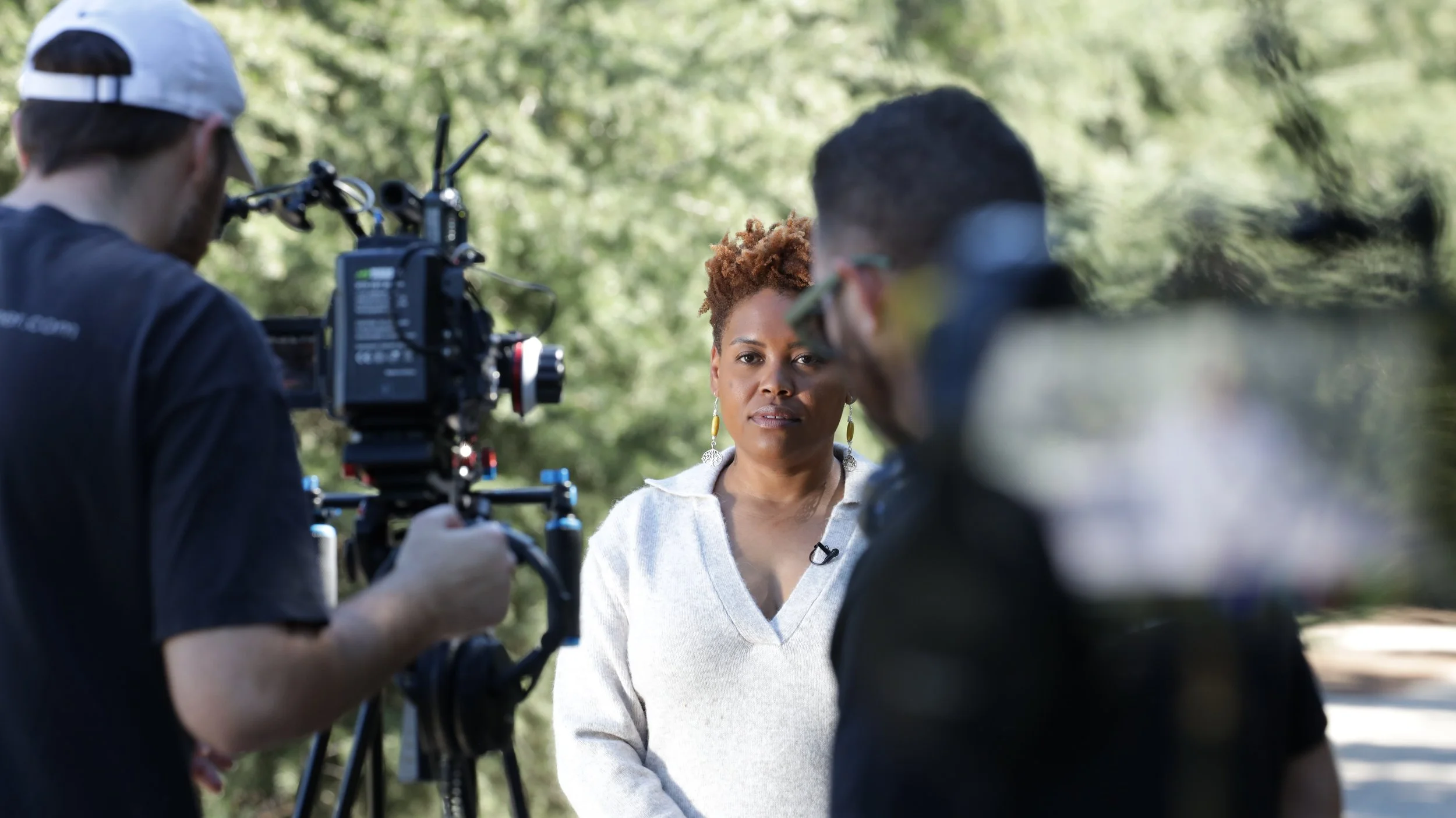 A woman being filmed outdoors with a camera crew, surrounded by trees.