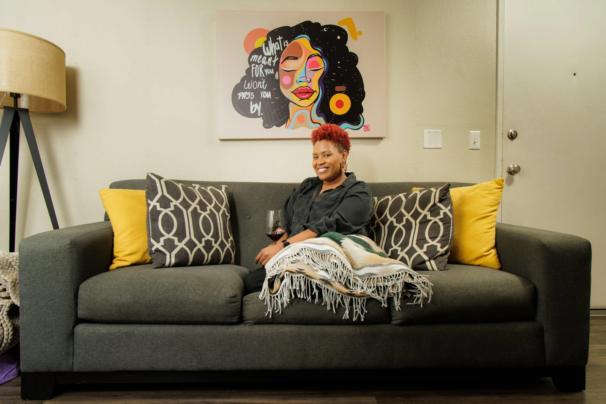 A woman with short, curly red hair sitting on a dark gray couch. She is holding a glass of red wine and smiling. The couch has yellow and patterned throw pillows. Behind her is a colorful painting of a woman's face with the text "What it is meant for you, you won't pass you by." The room has beige walls, a tall lamp on the left, and a closed white door on the right.