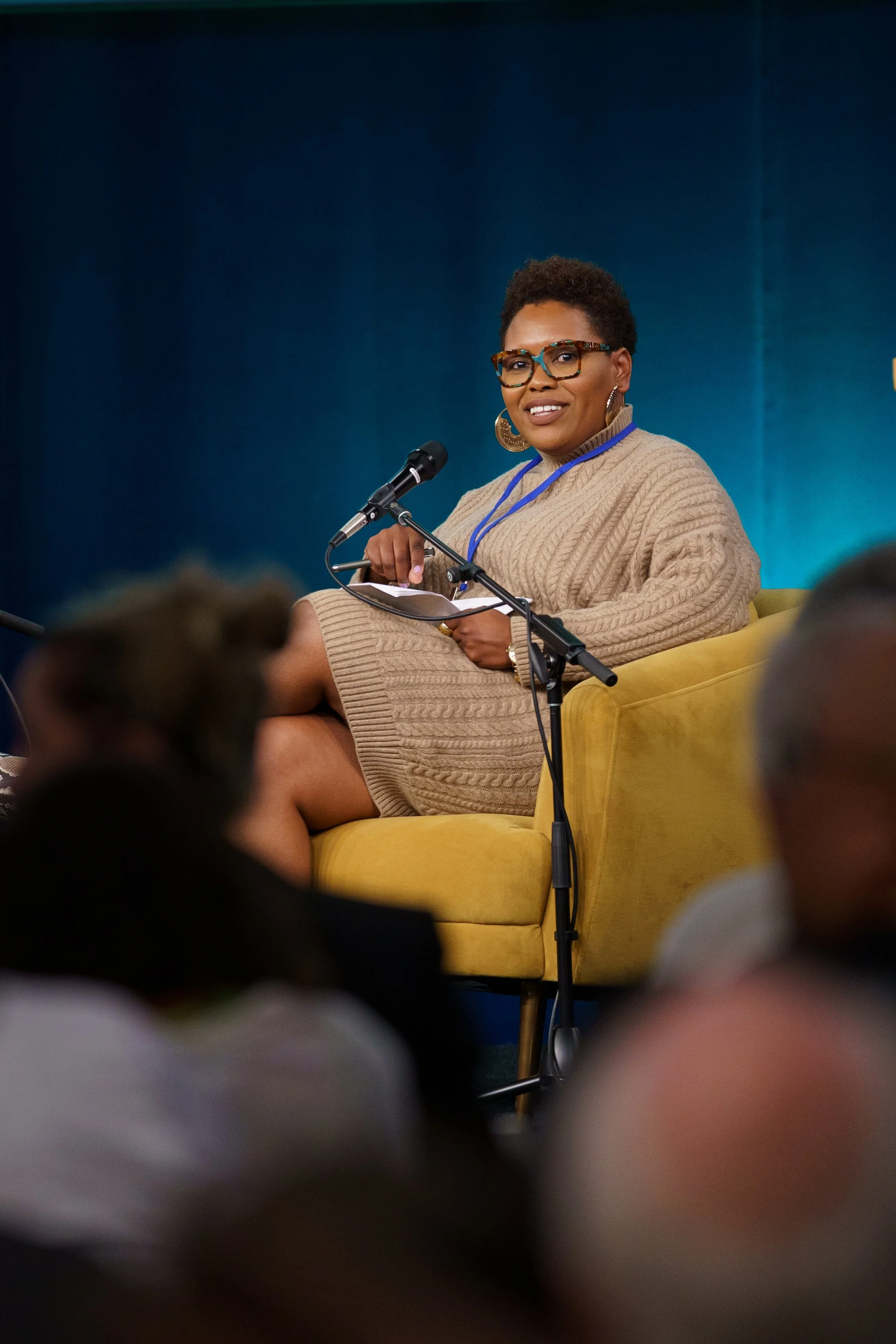 A woman with short curly hair, glasses, and earrings sitting on a yellow chair at a panel discussion or conference, smiling and holding a pen and paper, with a microphone in front of her against a blue background.