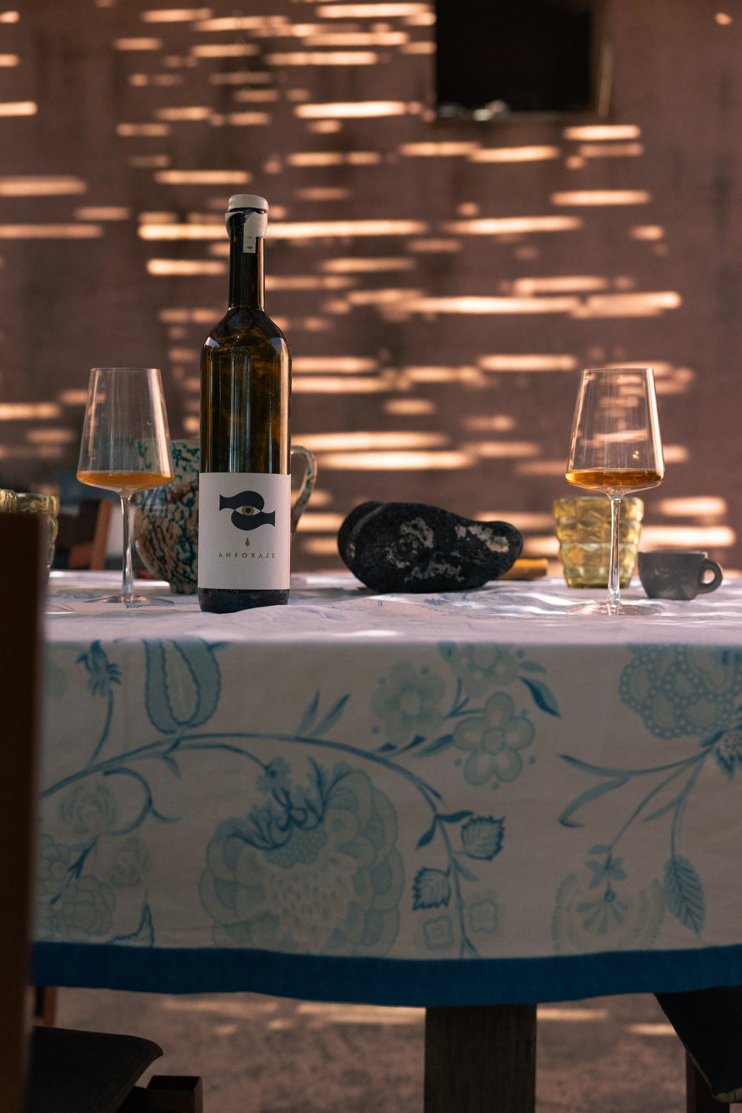 A wine bottle with a label that has a black fish and an eye design on a dining table, flanked by two wine glasses filled with a golden liquid, with a lace cloth covering the table and textured sunlight pattern on the background wall.