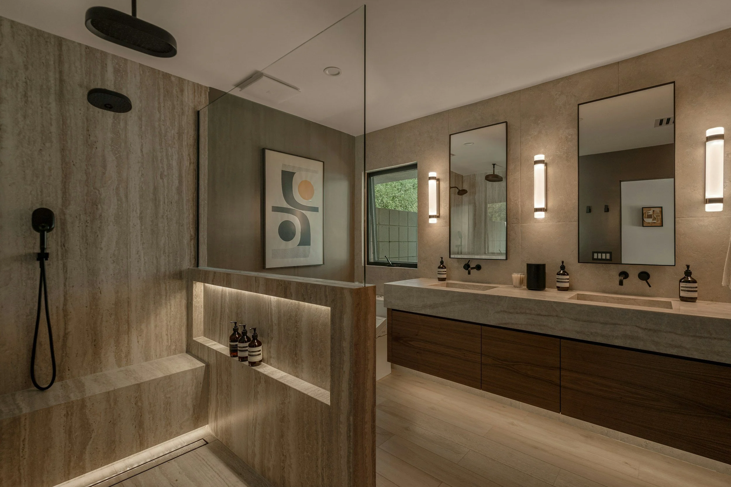 Modern bathroom with beige stone walls and flooring, featuring a dual-sink vanity with wooden cabinets, black and white striped soap dispensers, and three rectangular mirrors with vertical lights. A large window with greenery outside and a framed abstract artwork are visible. The shower area has a textured stone wall, a black handheld showerhead, and a built-in bench with bottles of toiletries placed on it.