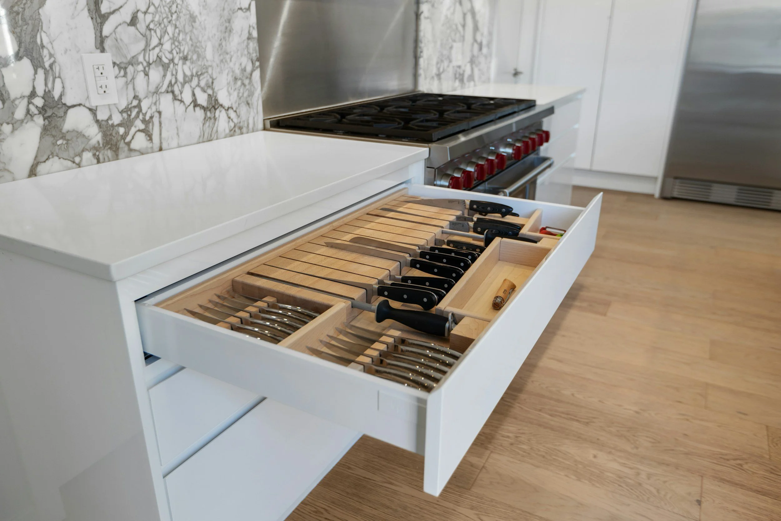 Open kitchen drawer filled with various kitchen knives and tools, with a marble backsplash and white countertops, in a modern kitchen setting.