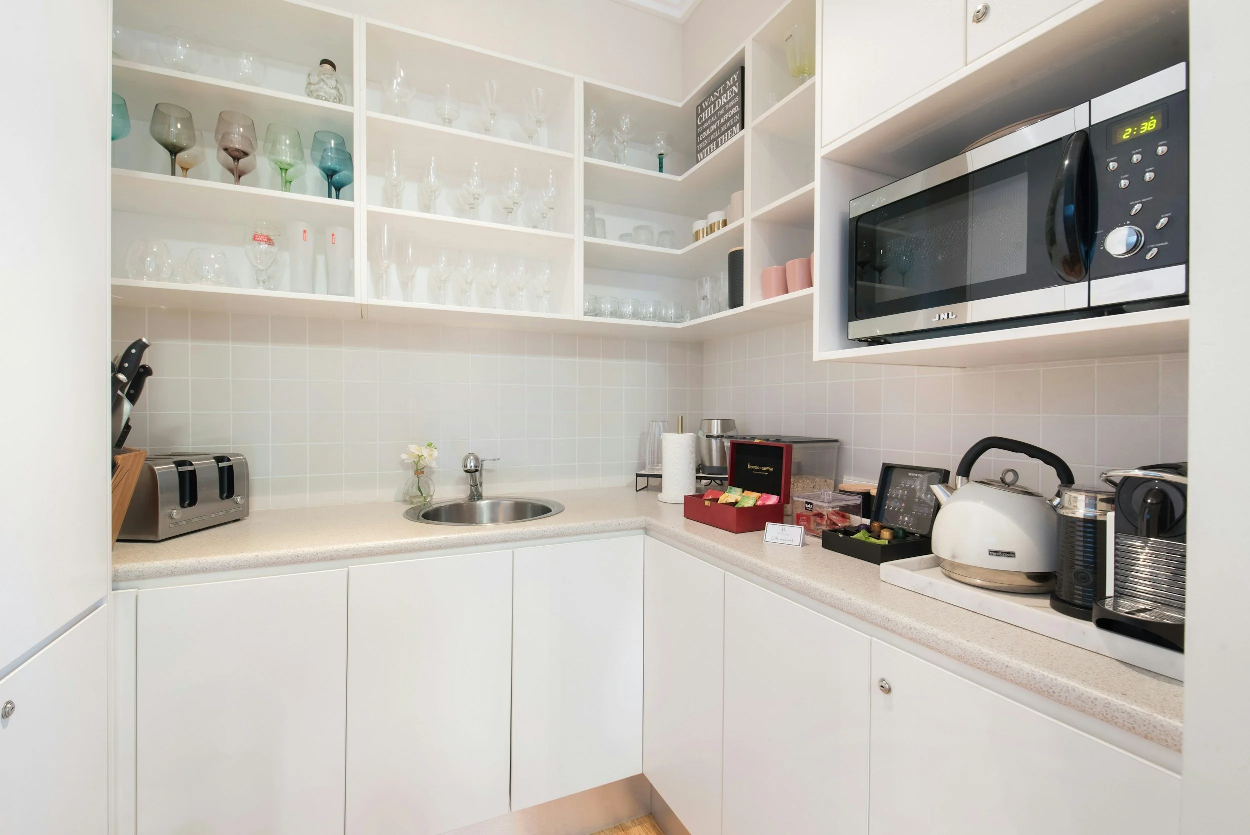 A kitchen with white cabinets, open shelving with glasses and mugs, a microwave oven, a toaster, a kettle, and various small kitchen appliances and items on the countertop.