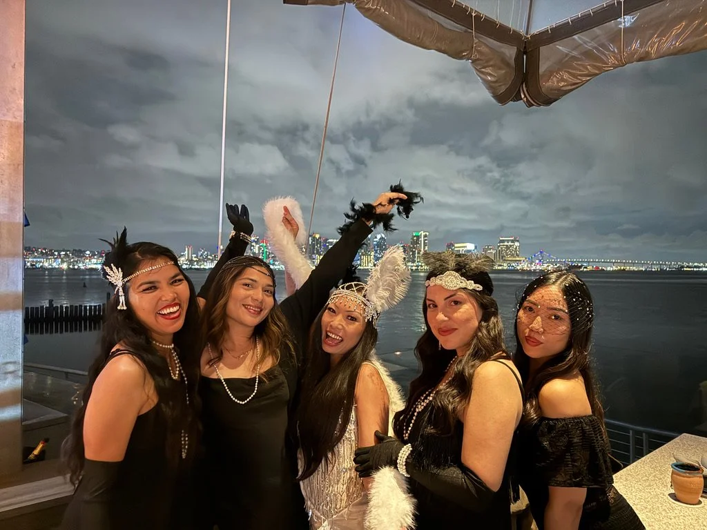 Five women dressed in vintage 1920s-style flapper costumes with headpieces and accessories, posing together on a rooftop balcony at night with a city skyline and river in the background.