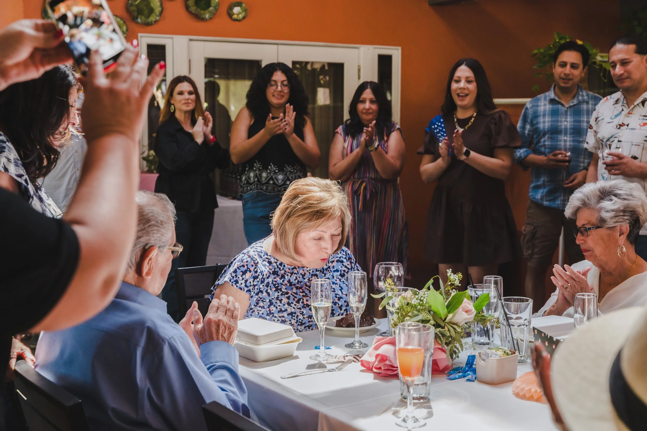 Group of people at a celebration, with women clapping and smiling, and elderly women seated at a dinner table with glasses of champagne and flower arrangements.