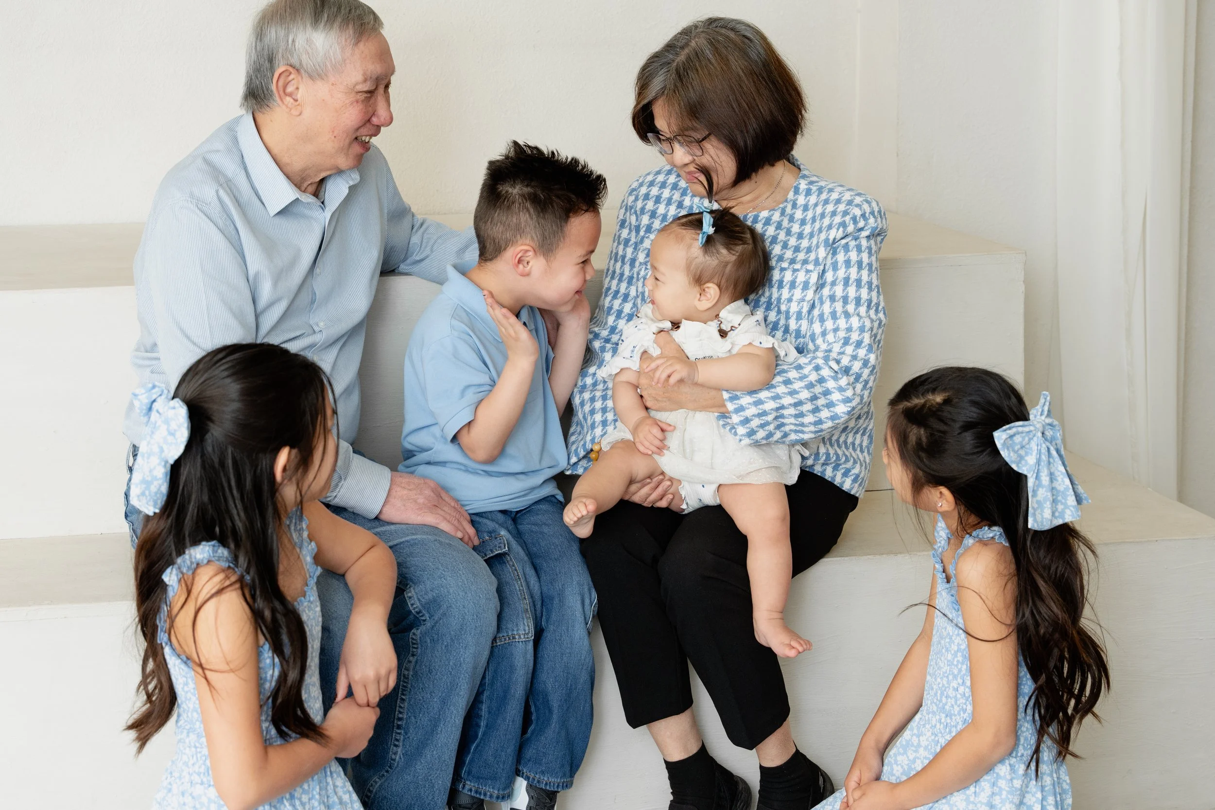 A family of six, including two elderly people, a woman, and three young girls with hairbows, sitting on stairs engaging with a baby girl held by the woman.