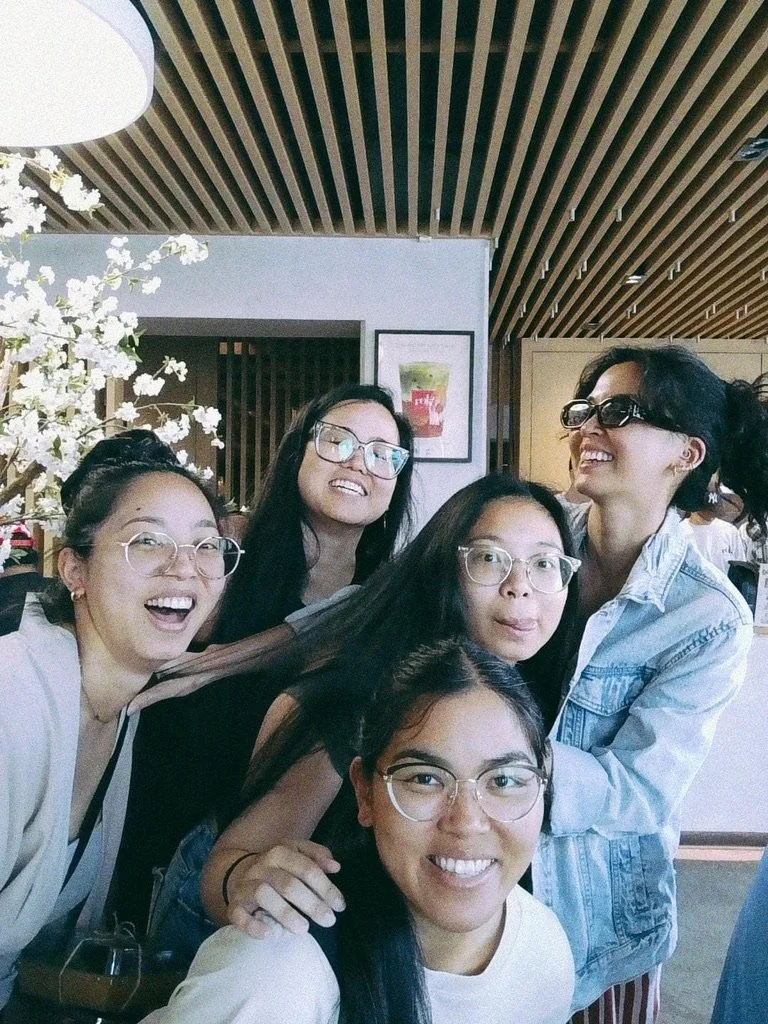 Group of five women smiling and posing together indoors, with wooden ceiling and framed pictures on the wall in the background.