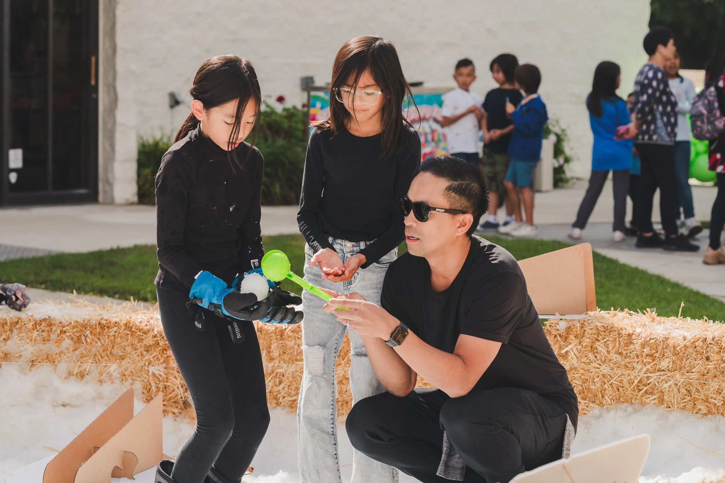 A man with sunglasses kneeling and talking to two young girls holding a toy robot with a green shovel, outdoors at an event with other children in the background.