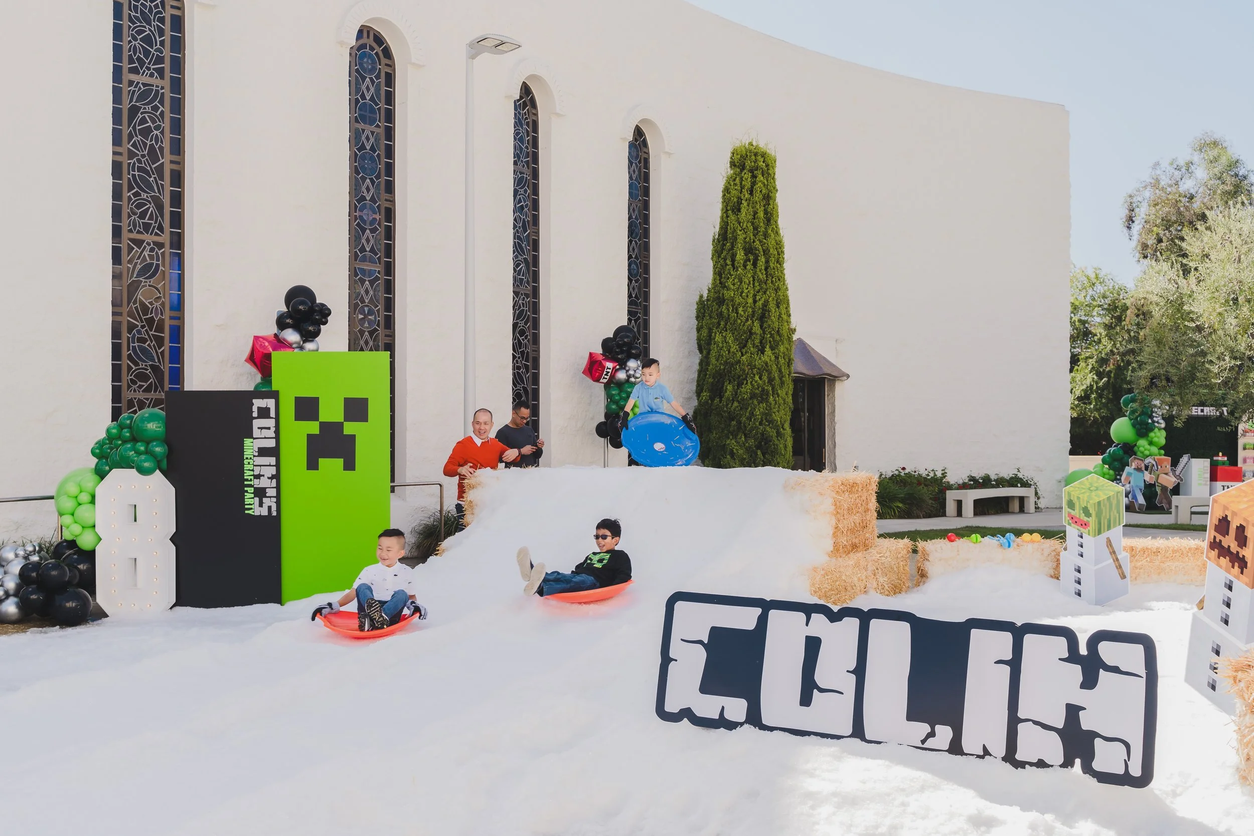 Children playing on a snow-themed playground with hay bales, Minecraft-themed decorations, and a church-like white building with tall stained glass windows in the background.