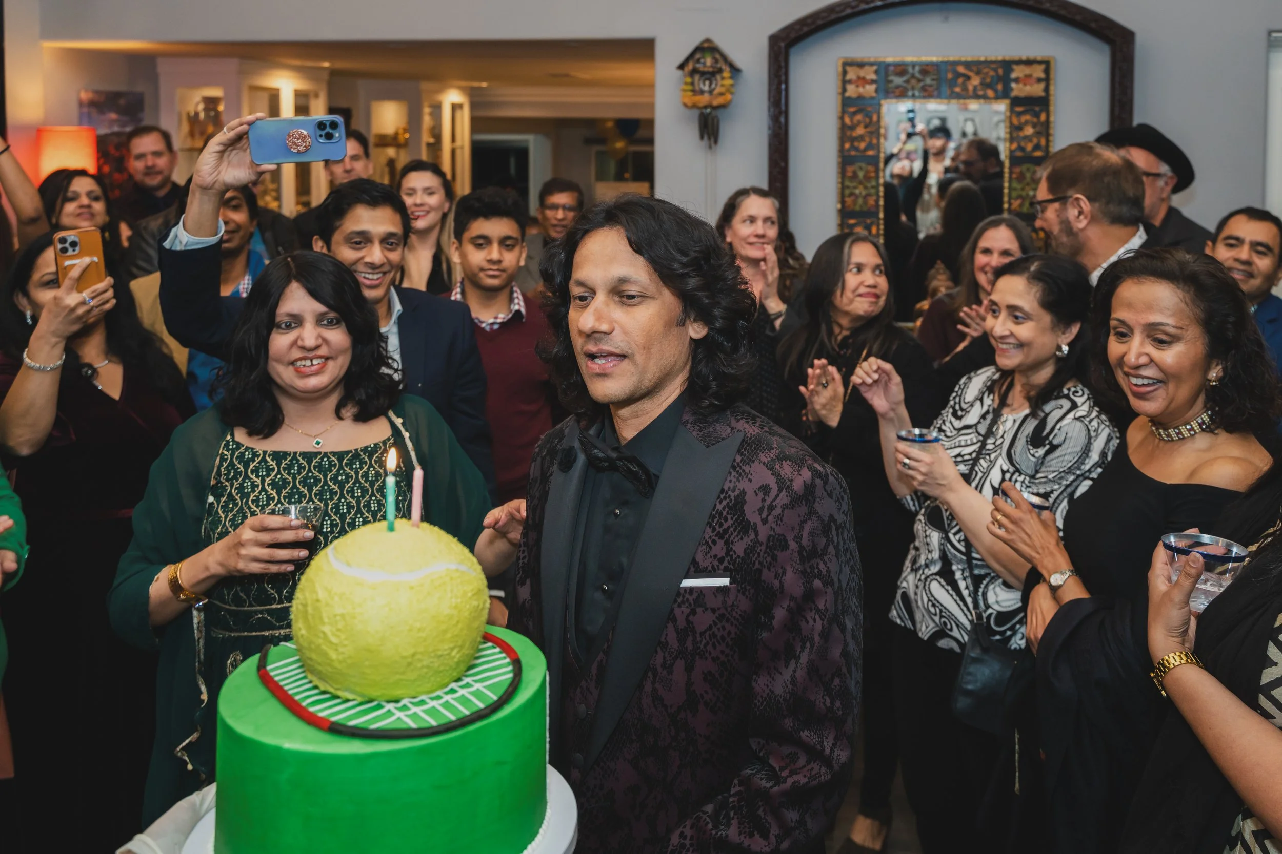 A group of people celebrating a birthday at a party. A man with long black hair and a suit stands in front of a yellow birthday cake with a lit candle. Many guests are smiling and taking photos, some holding drinks. The setting is indoors with decora