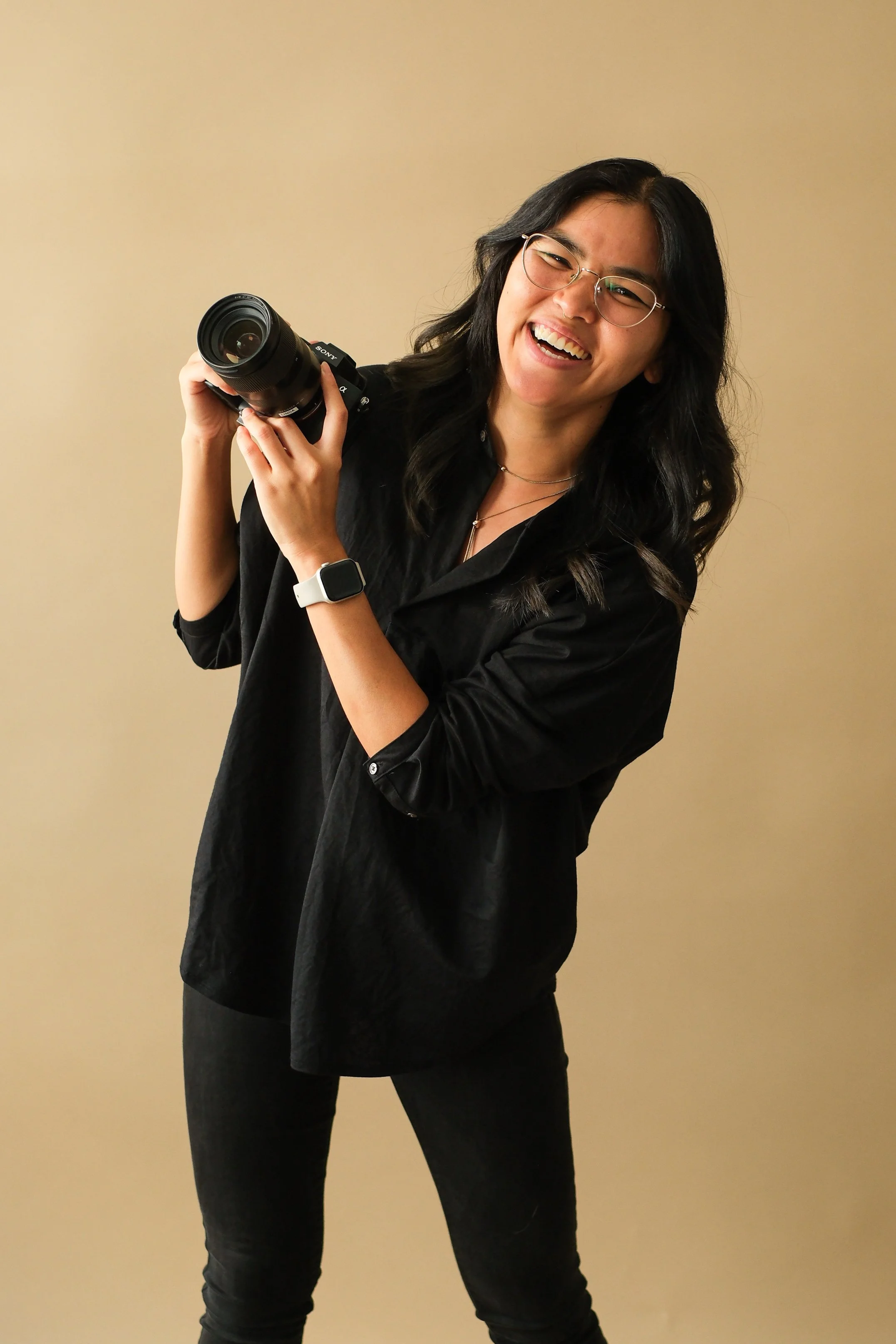 A woman with glasses and dark hair smiling, holding a camera, wearing a black outfit, standing against a plain beige wall.