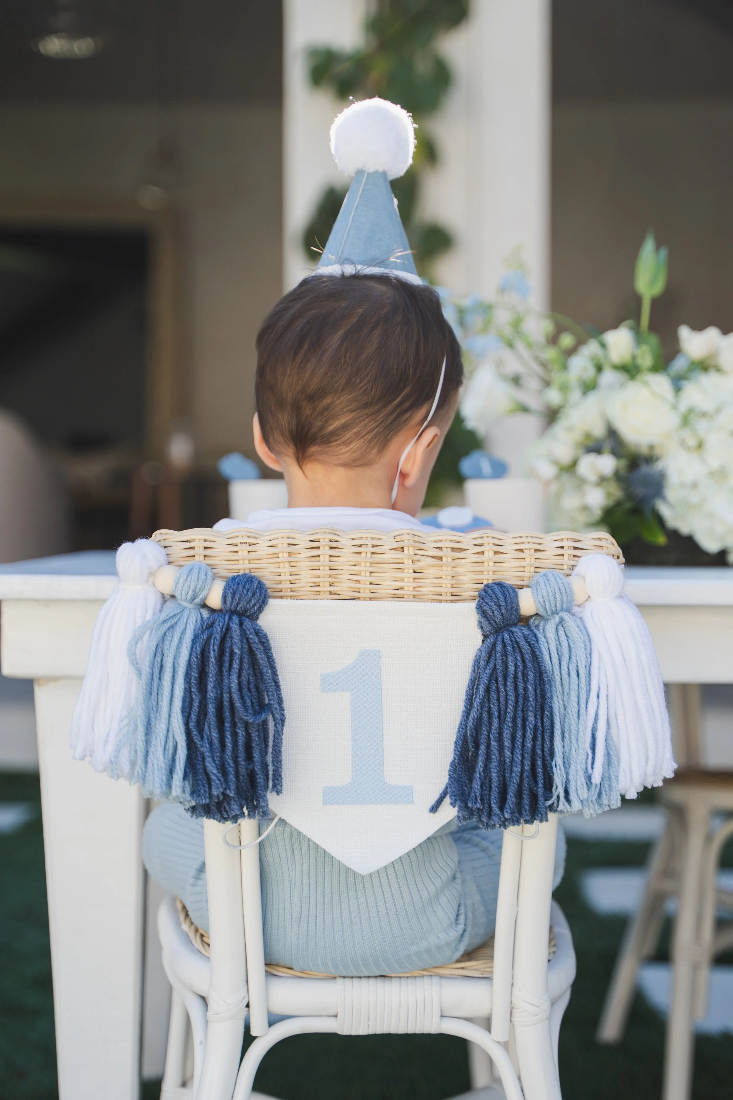 Back of a young child wearing a blue and white party hat, sitting in a white chair in front of a decorated table with white flowers. The chair has a sign with the number 1.