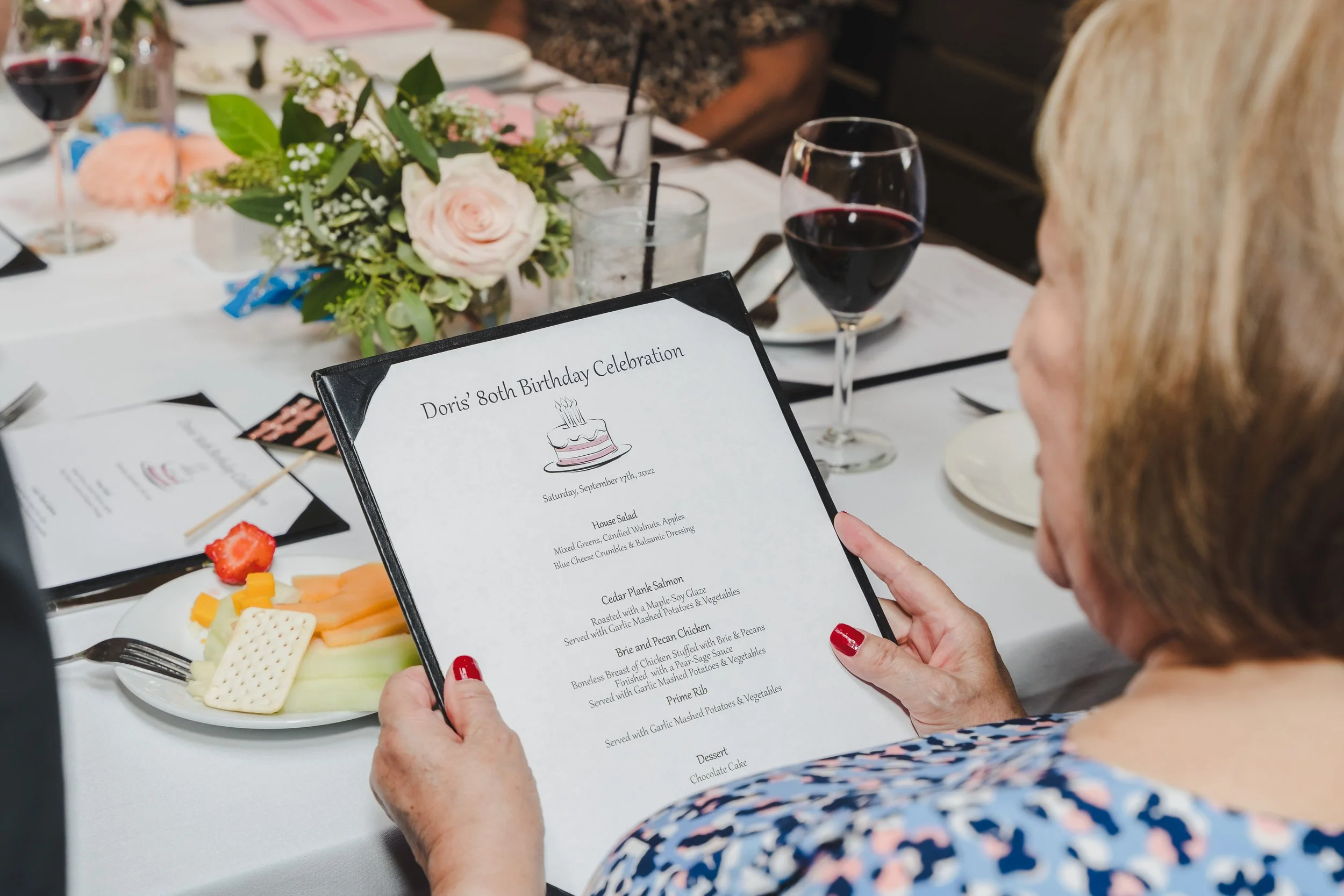 A woman holding a birthday dinner menu at Doris' 80th birthday celebration. The table features a floral centerpiece, wine glasses, and various food items.