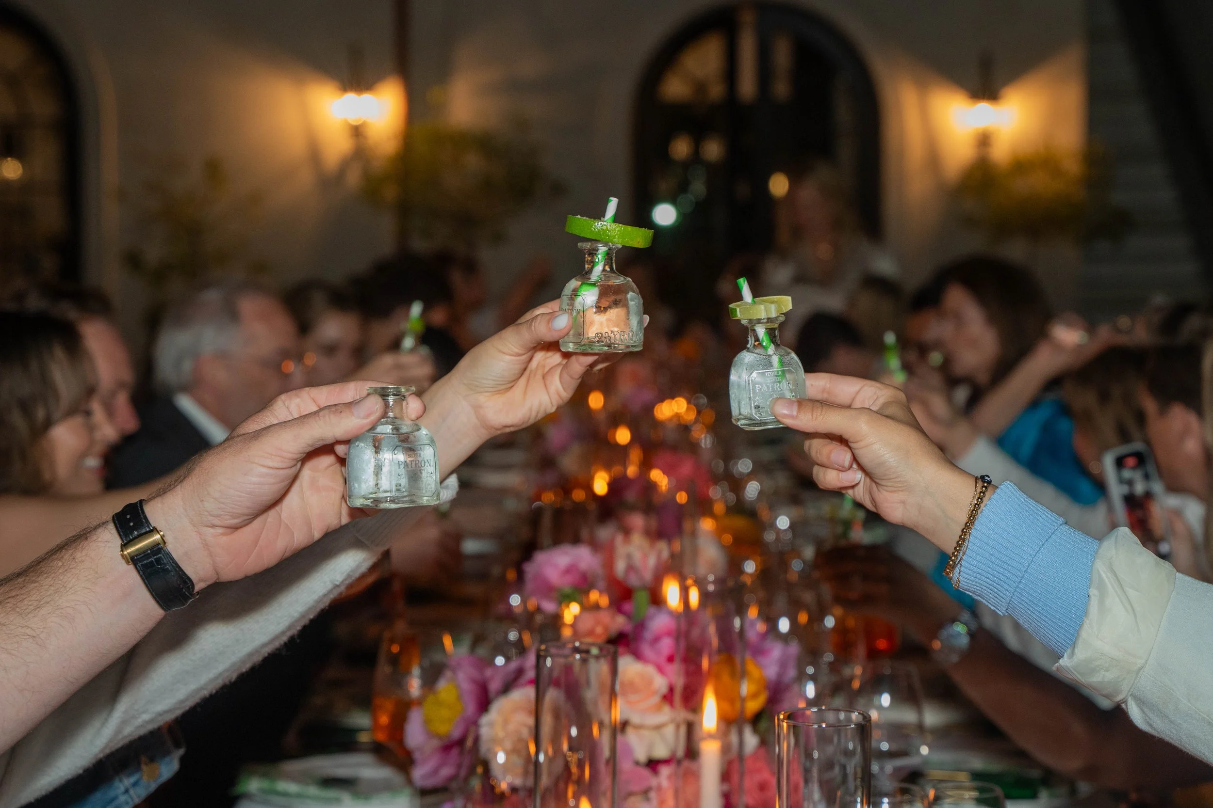 Guests raising shot glasses filled with clear liquor at a crowded dinner party.
