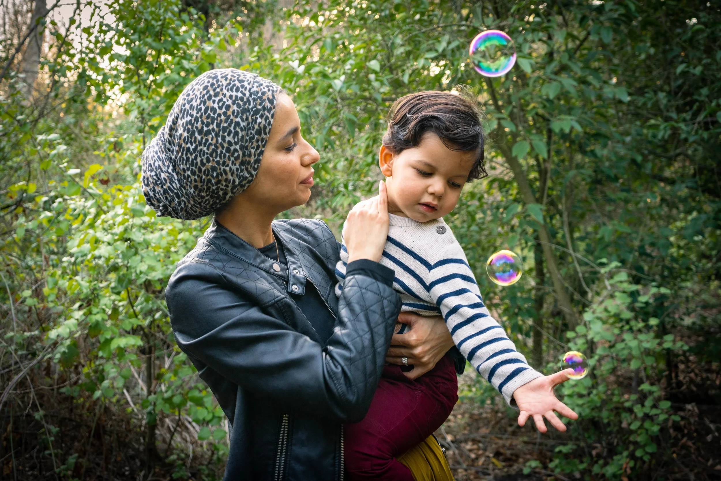 A woman wearing a leopard print headscarf and black leather jacket is holding a young boy in a striped sweater as they watch soap bubbles floating in the air outdoors surrounded by green trees and bushes.