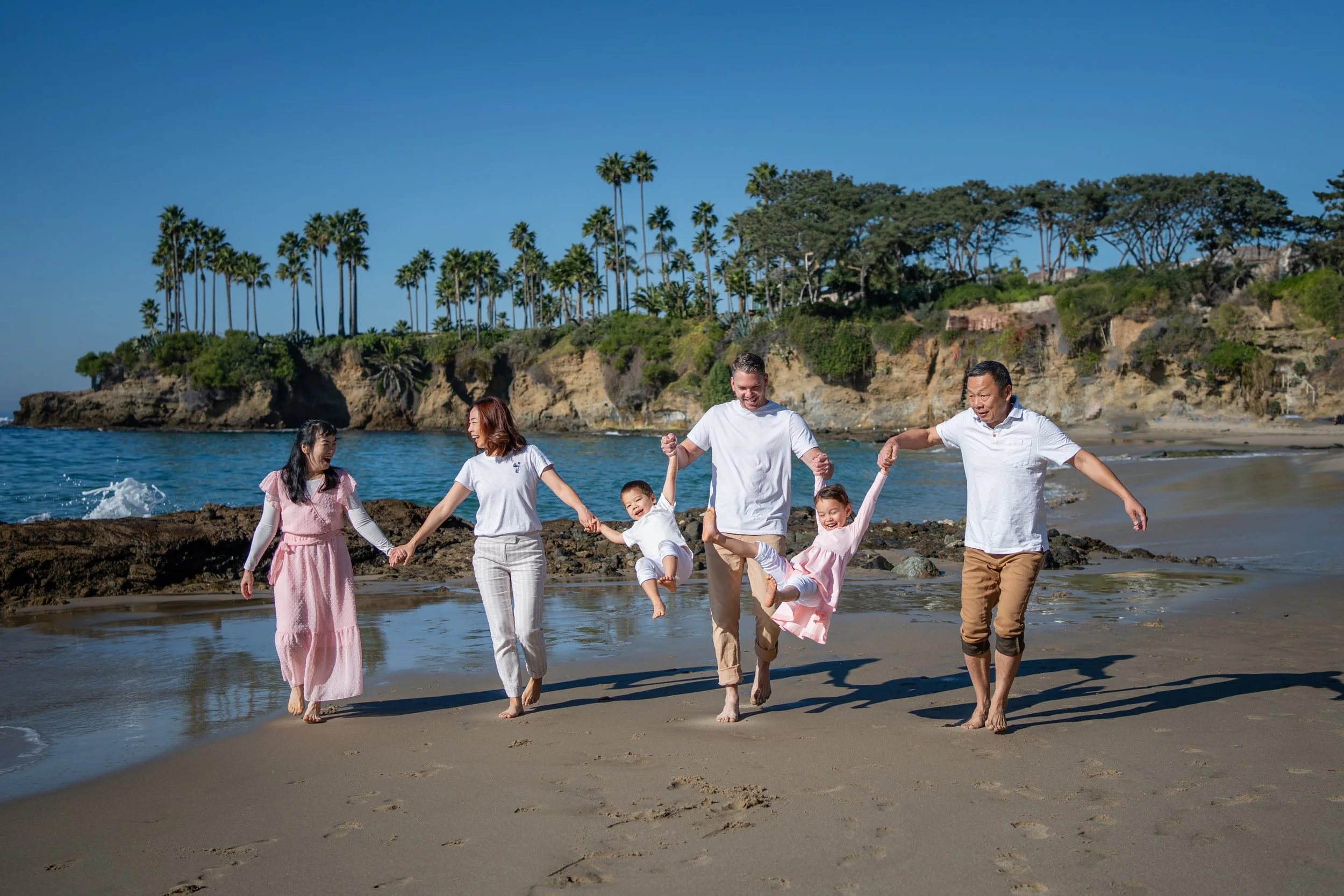 Family of five holding hands and playing on the beach with cliffs and palm trees in the background.