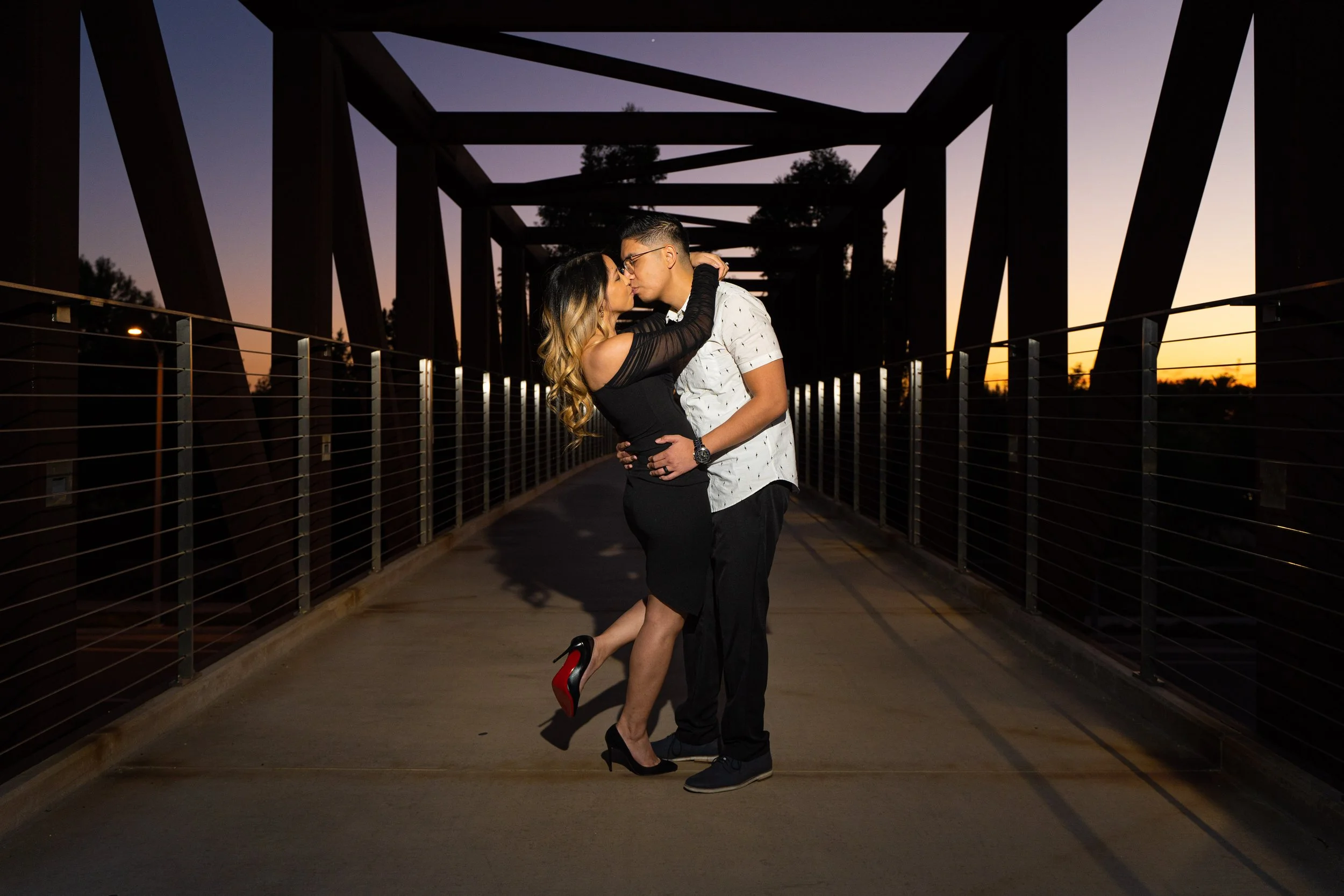 A couple is embracing on a bridge during sunset, resembling a romantic moment, with one person lifting the other.