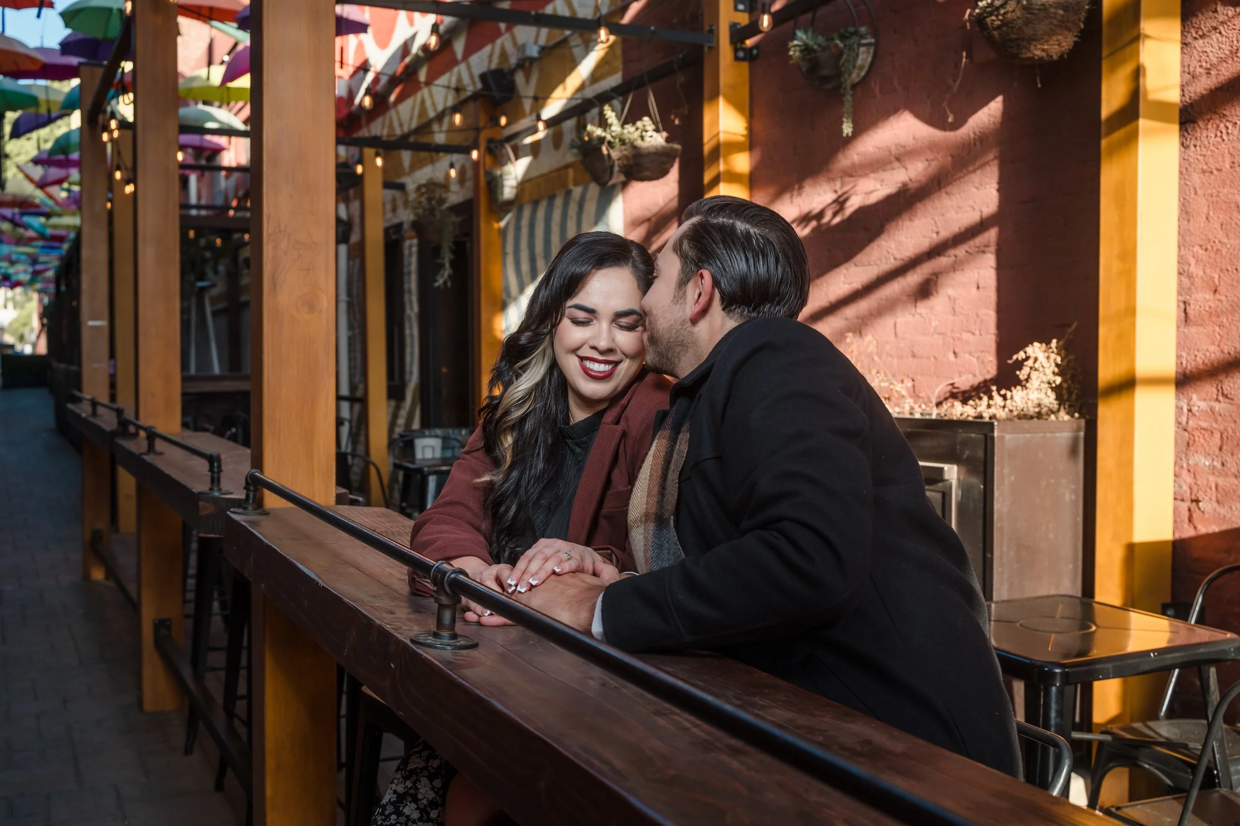 A couple sitting at a wooden table outdoors, enjoying a romantic moment with a kiss on the forehead. The woman has long dark hair and is smiling, while the man has slicked-back hair and is leaning in to kiss her. Colorful umbrellas and a decorated ou