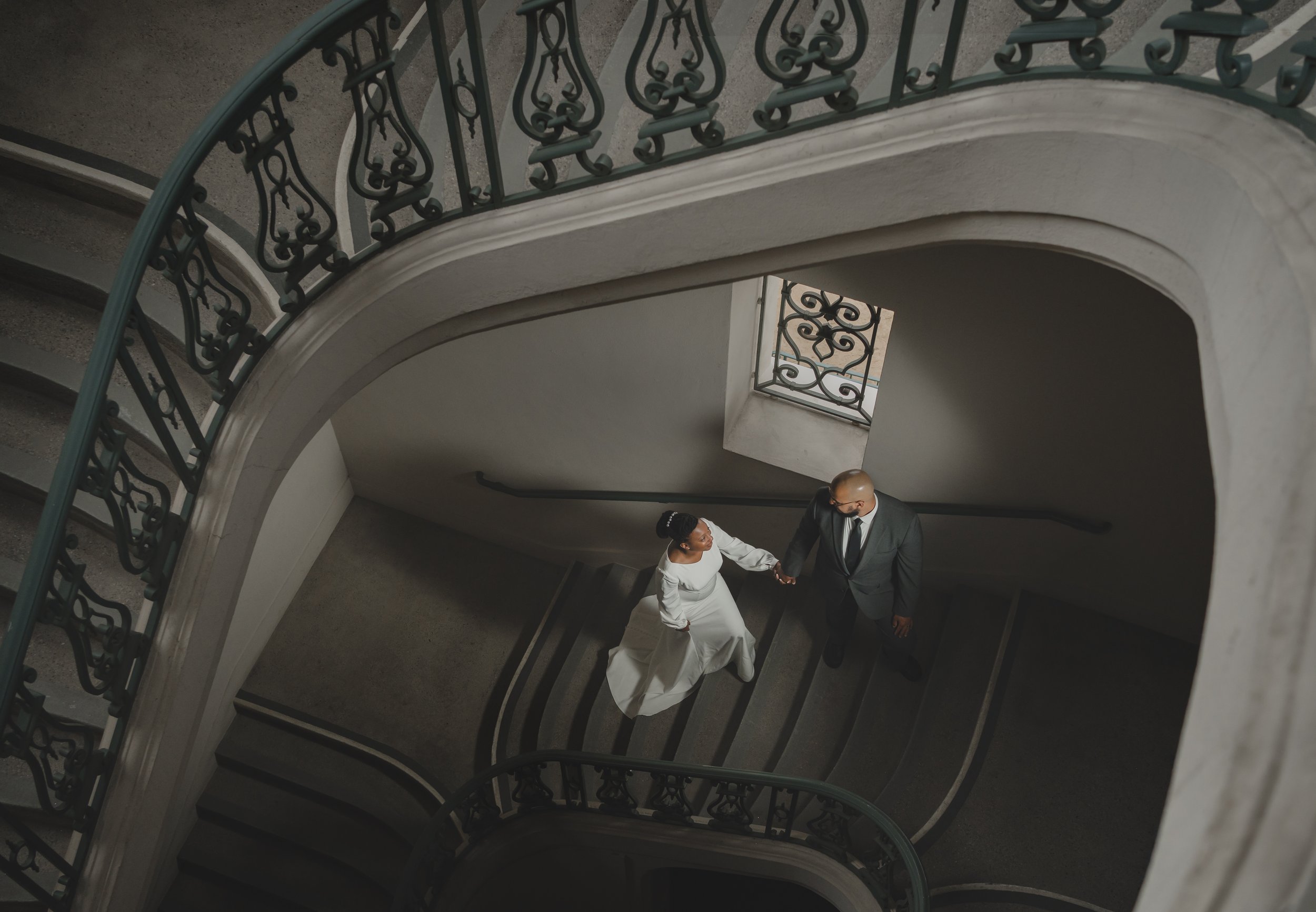 A bride and groom standing on a spiral staircase, holding hands, with ornate black railings and a window in the background.