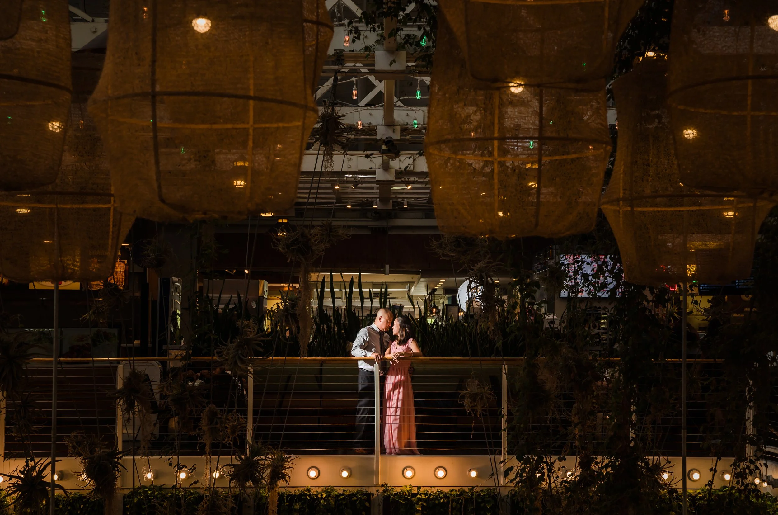 A man and woman standing close together on an indoor balcony, engaged in conversation. The space is decorated with large hanging lanterns and string lights, with indoor plants and a modern interior design in the background.