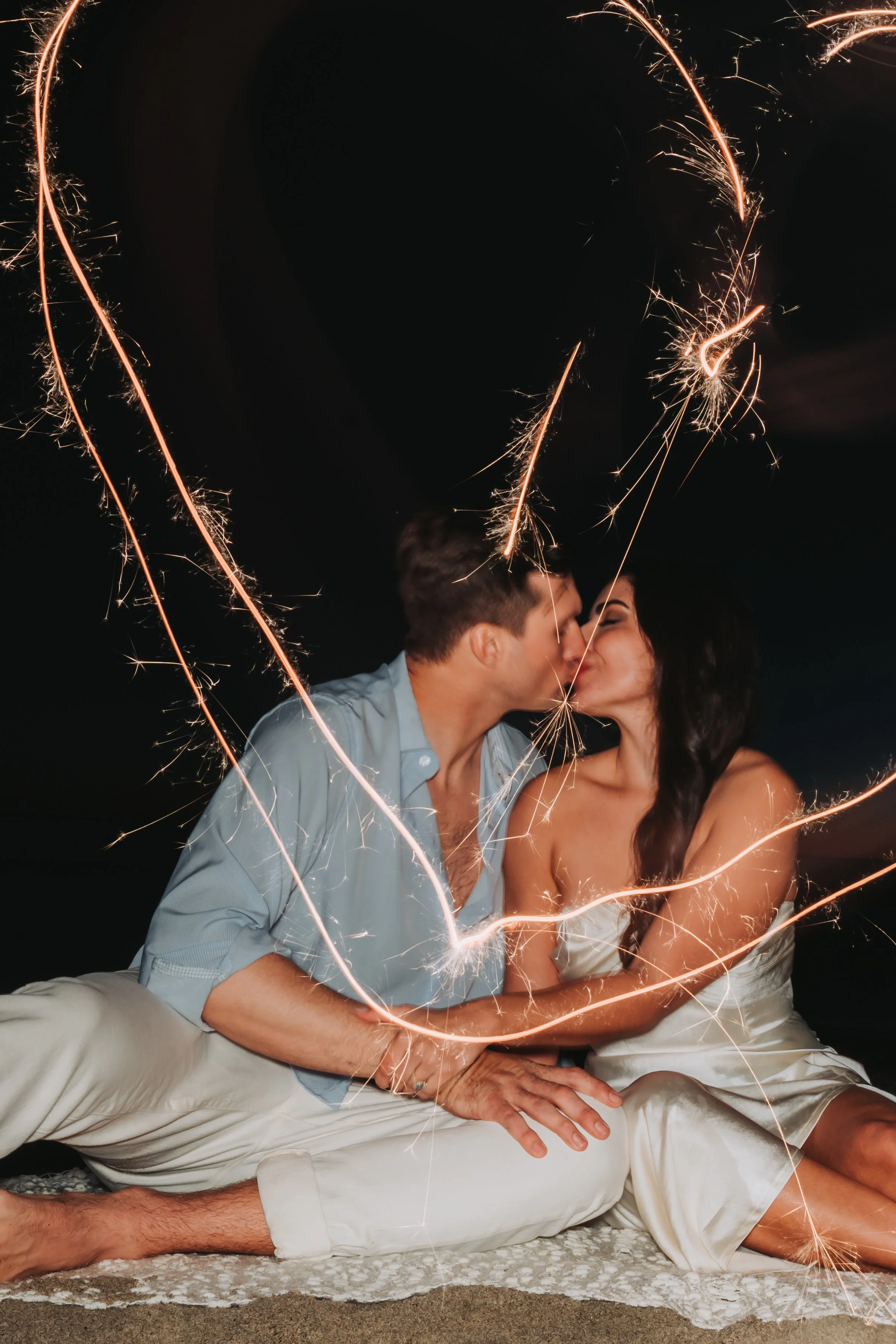 Couple sharing a kiss behind sparklers at night, sitting on a blanket, with a dark background.