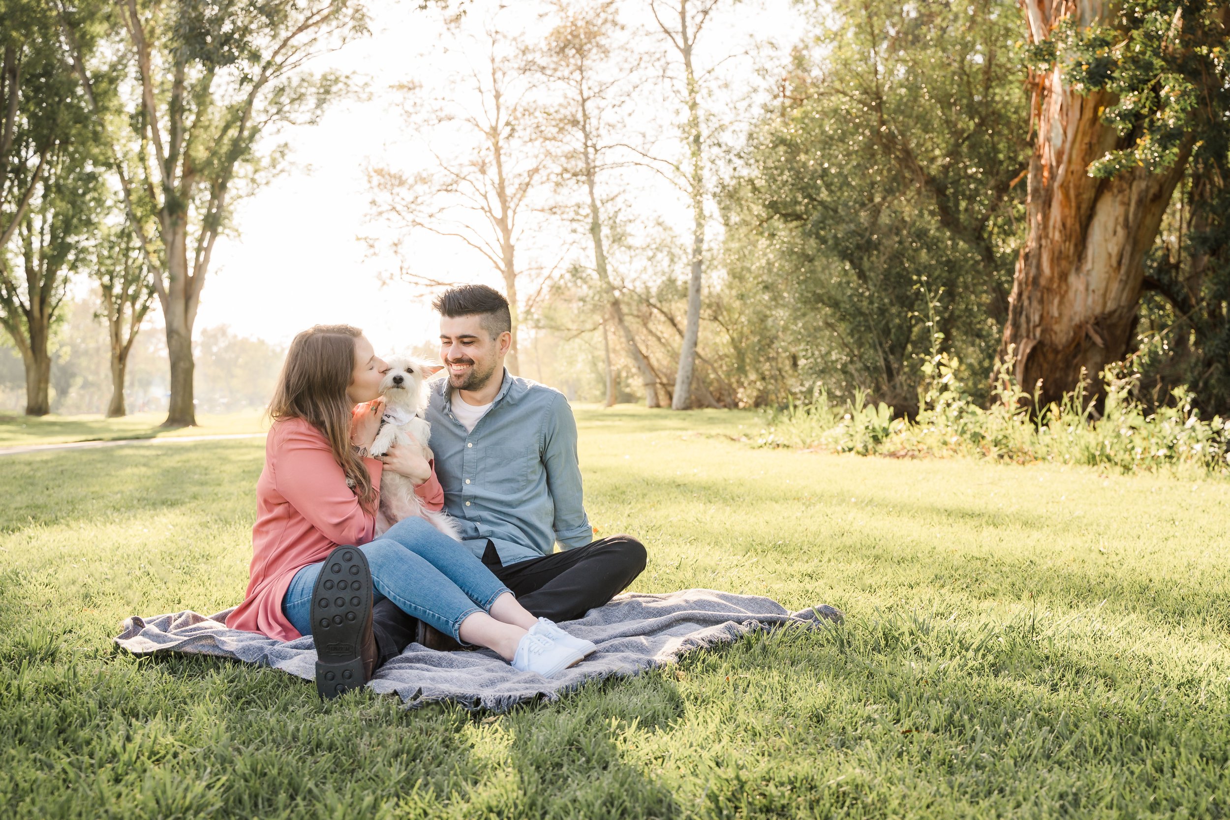A woman, a man, and a small white dog sitting together on a blanket in a park, with trees and sunlight in the background.