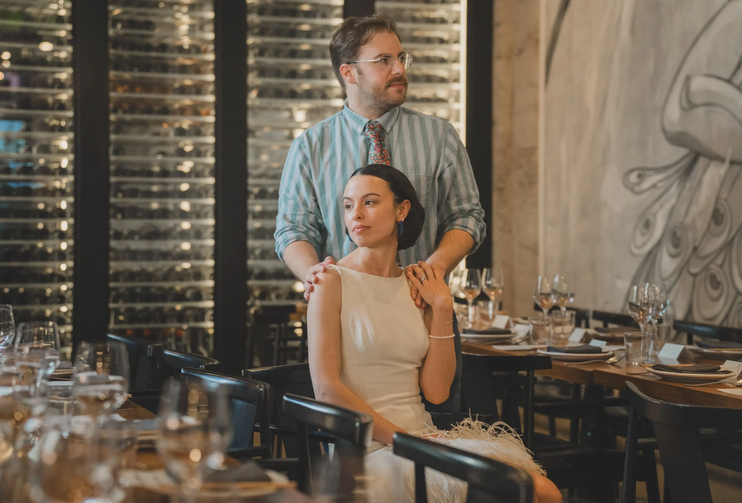 A man and a woman are inside a restaurant; the man stands behind the seated woman, gently placing his hands on her shoulders. The woman wears a cream-colored dress, and the man wears glasses, a striped shirt, and a colorful tie. The restaurant is dec