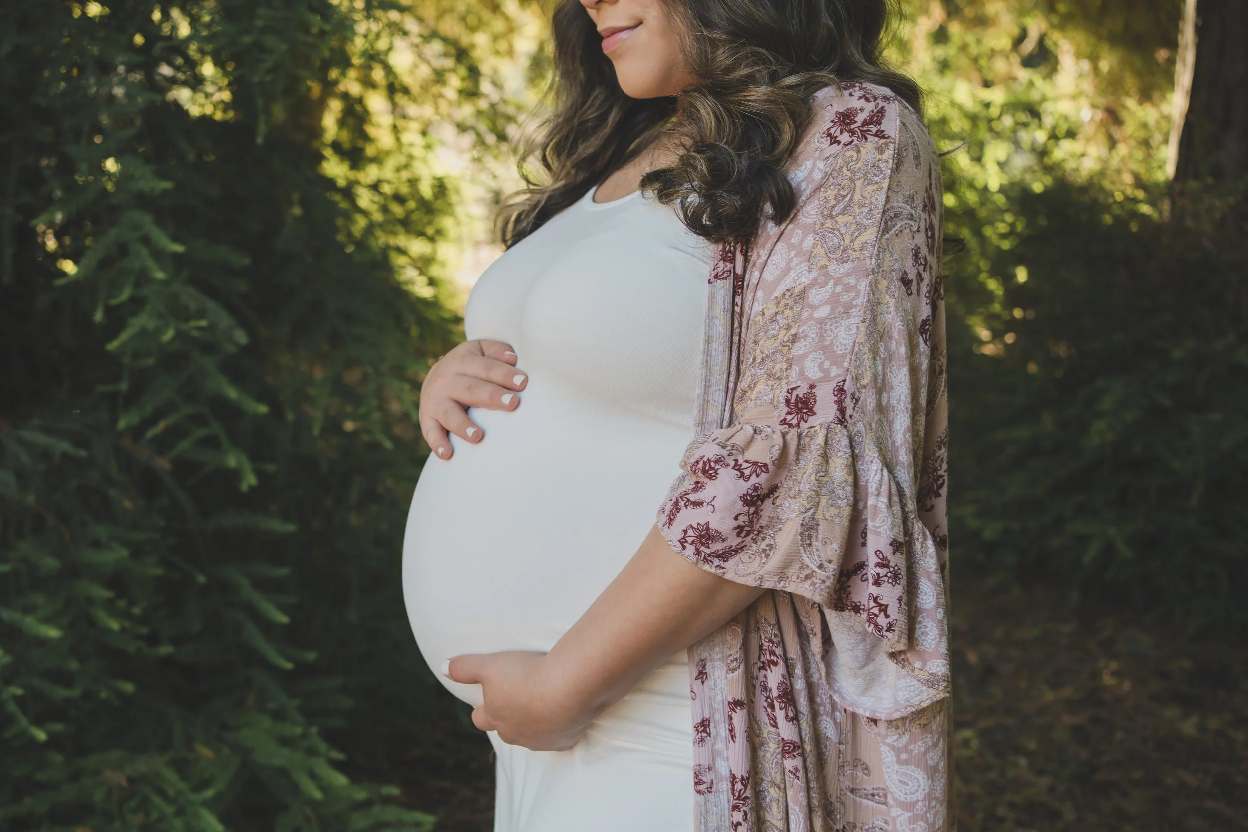A pregnant woman with long dark hair, wearing a white top and a pink floral shawl, standing outdoors in a green, leafy environment, gently holding her rounded belly.