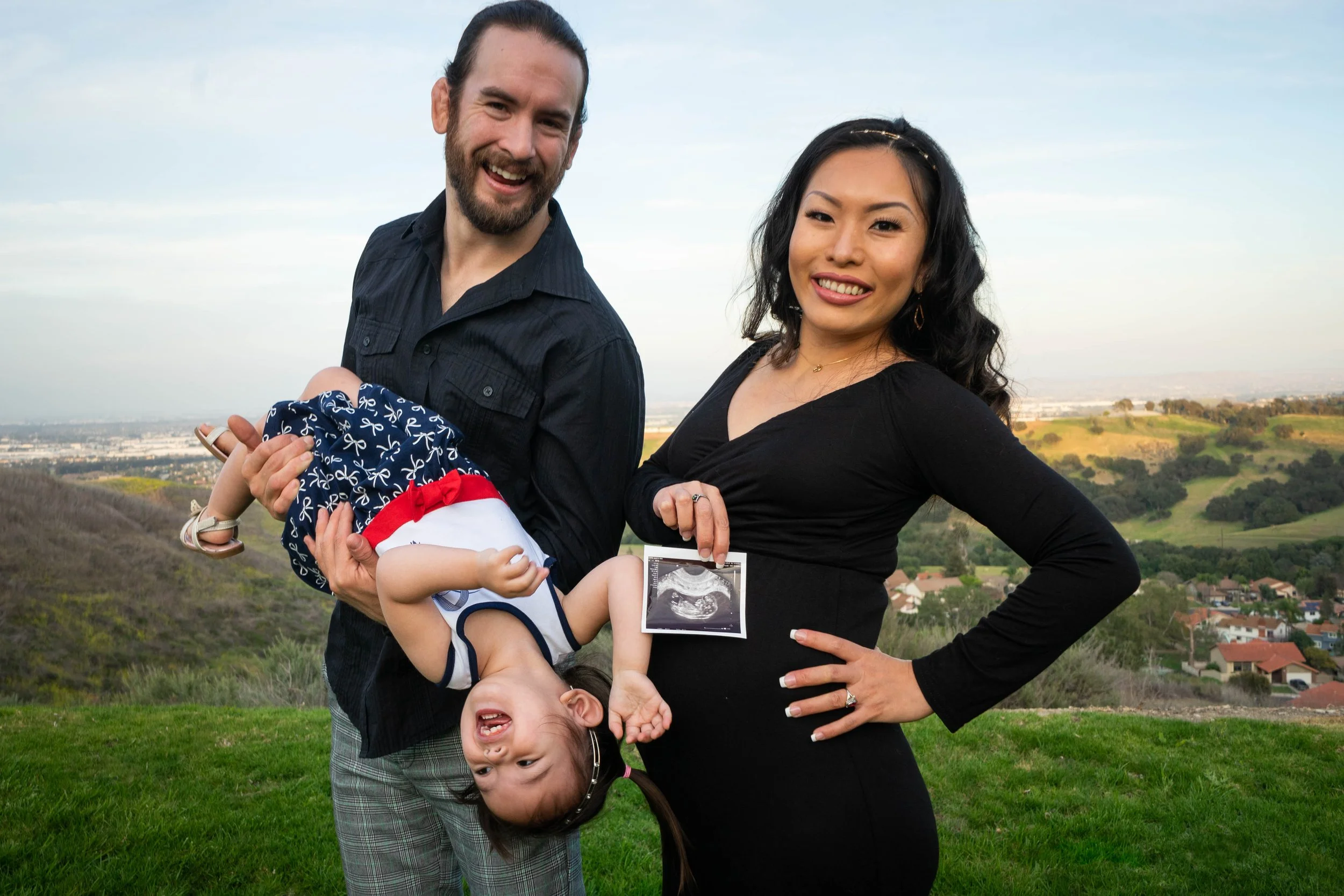 Family celebrating pregnancy outdoors, holding ultrasound image, with rolling hills and houses in background.