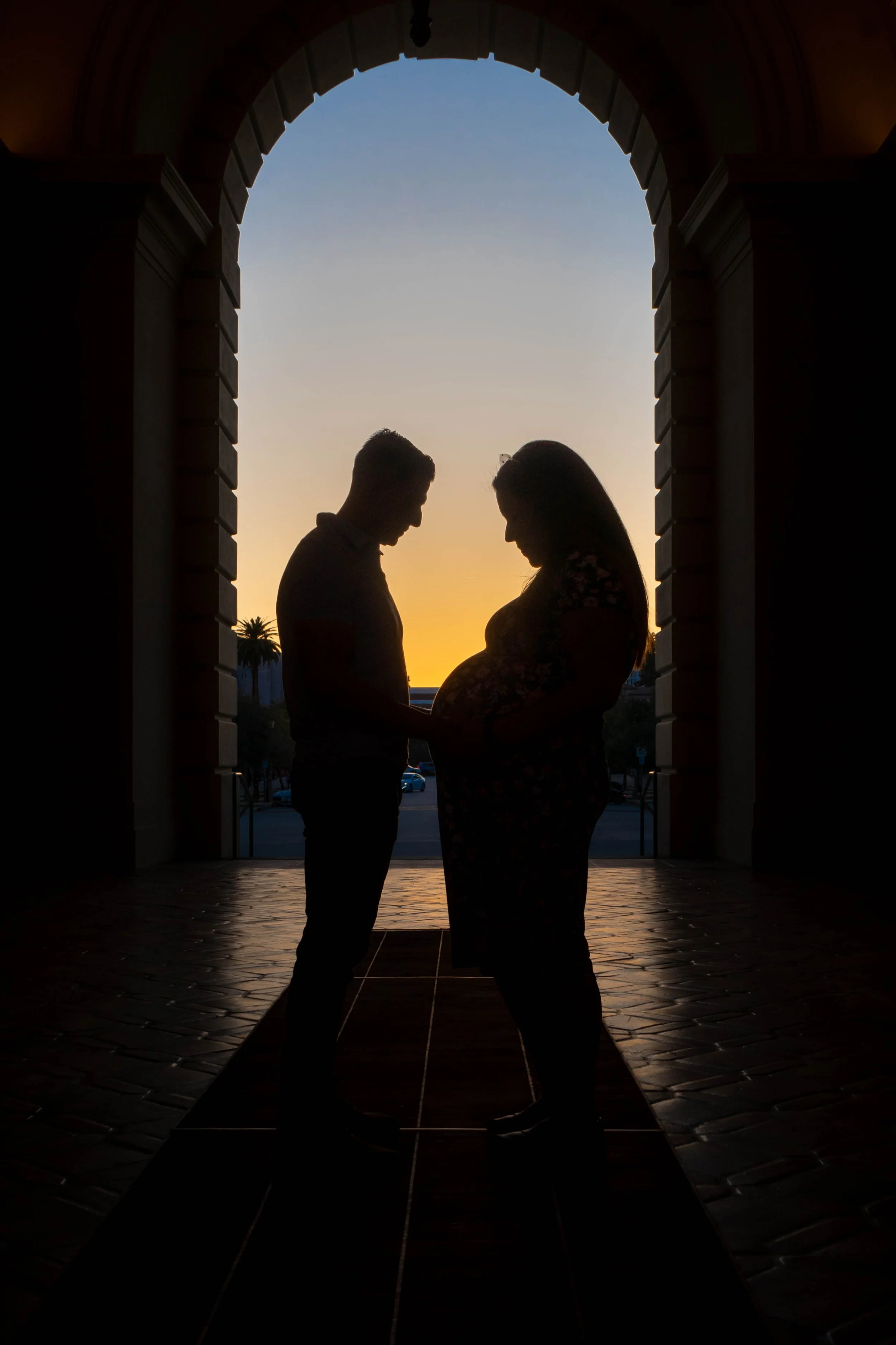 A silhouette of a pregnant woman and a man, possibly her partner, holding hands and touching her belly under an archway during sunset, with palm trees and a parked car in the background.